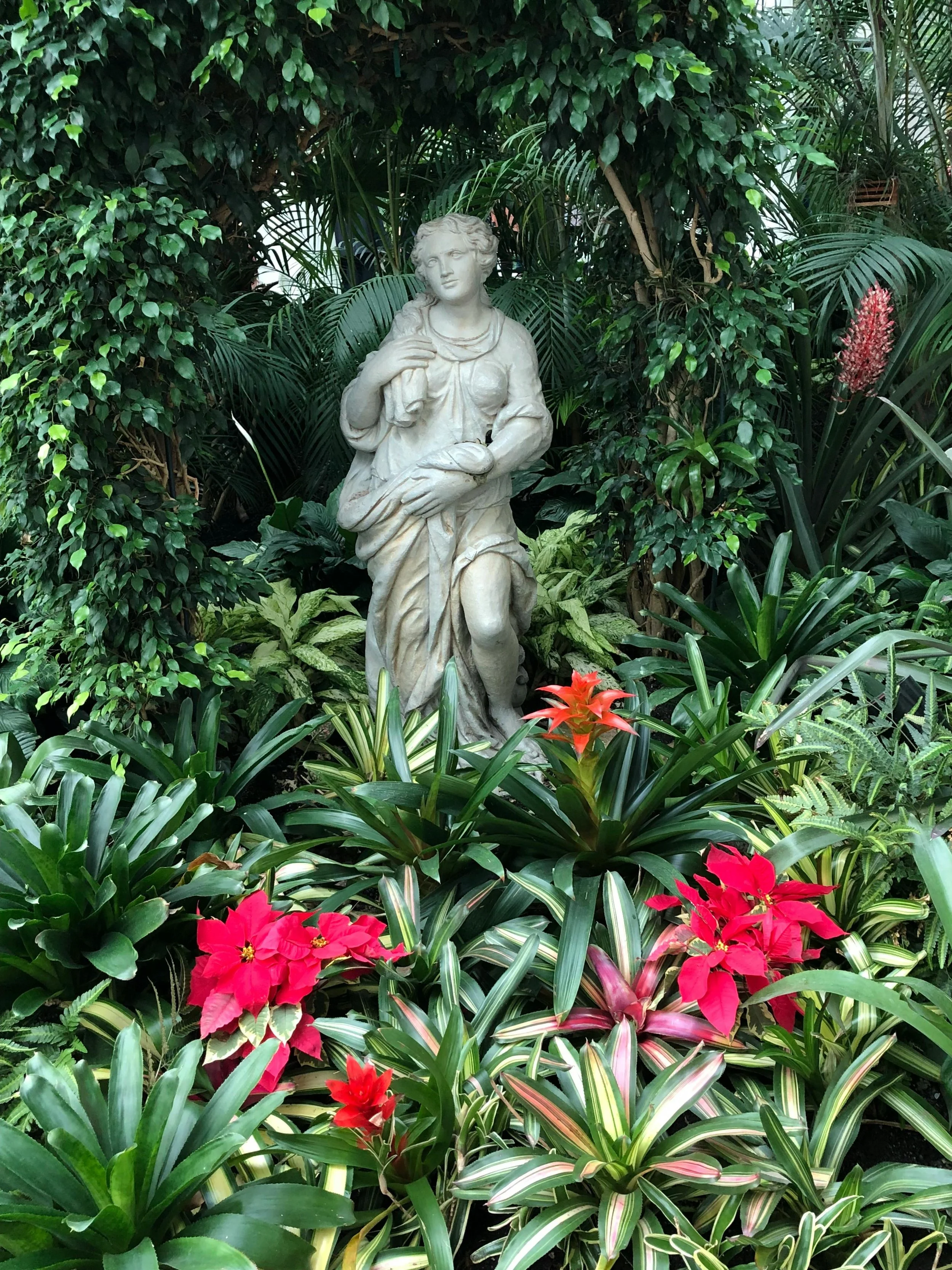 A white marble statue of a woman in a garden surrounded by lush green plants, pink and red flowers.