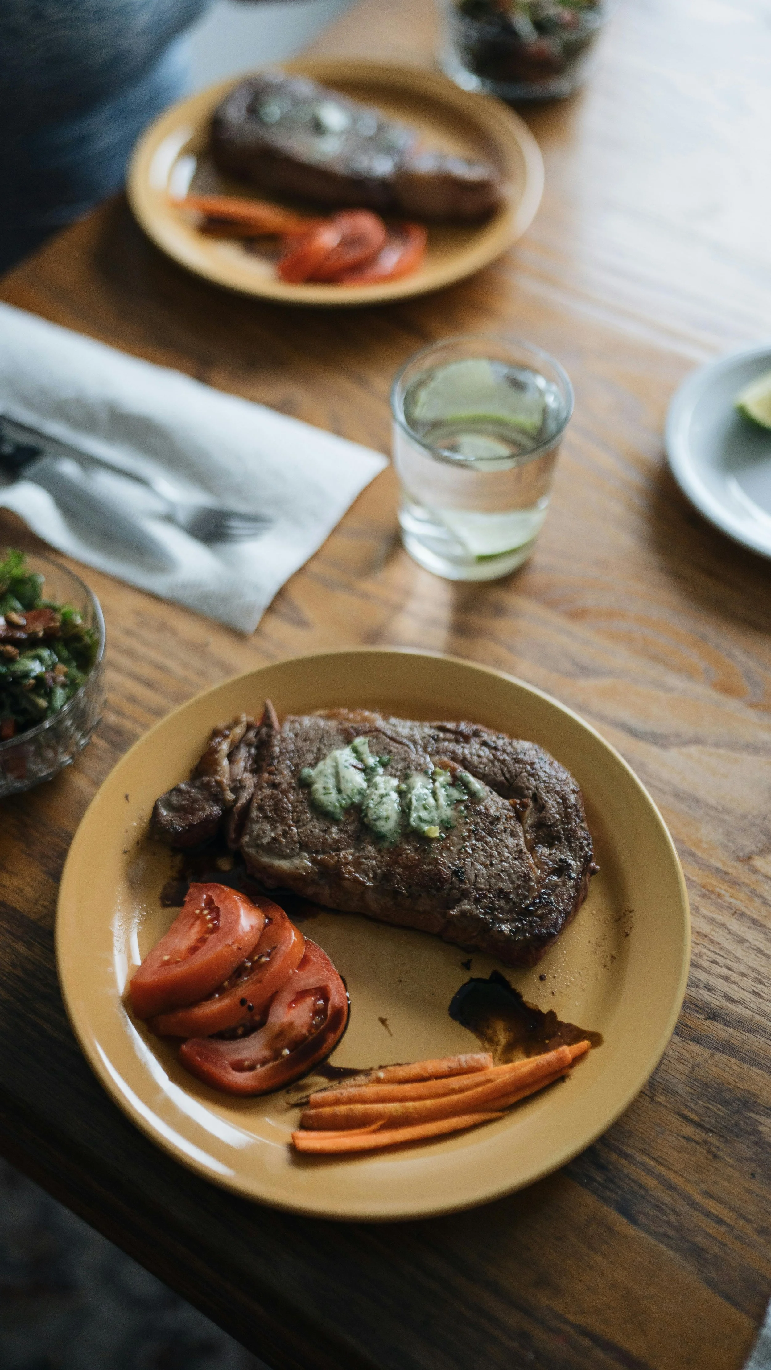 A plate with a cooked steak topped with butter, sliced tomatoes, shredded carrots, and a small bowl of salad on the side, on a wooden table with a glass of water nearby.