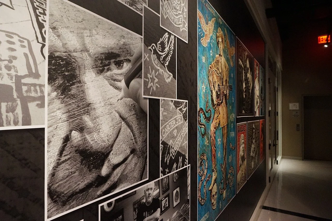 A hallway display wall with various art posters, including a black-and-white photograph of a person's face with a marker, tattoo designs, a singer with a guitar, and other artistic images.