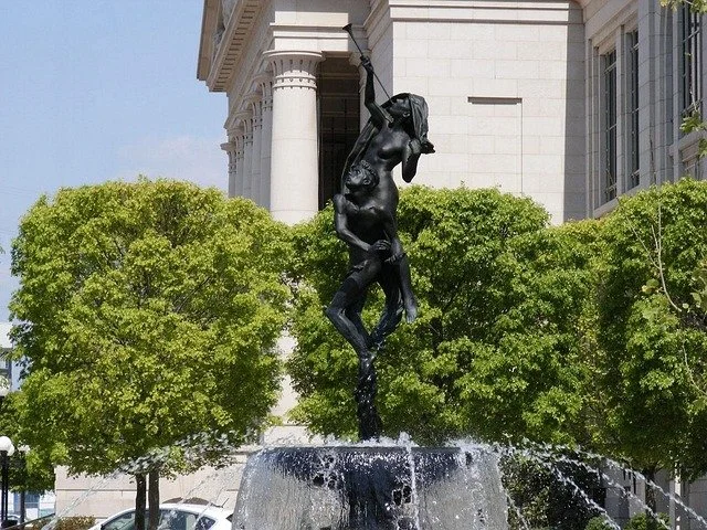 Black sculpture of a man hanging from a ledge, positioned above a fountain, with trees and a building in the background.