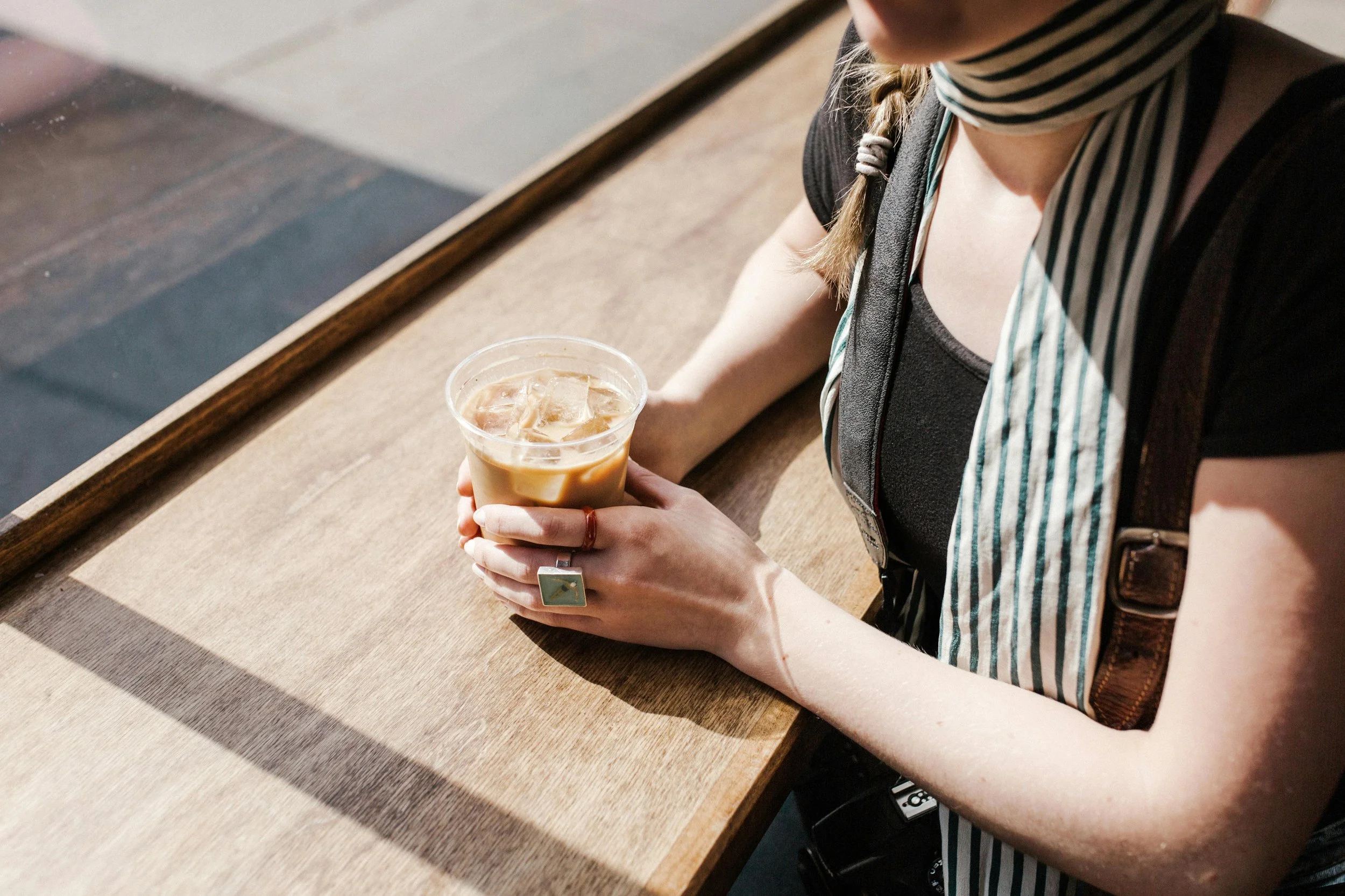 Person holding iced coffee with ice cubes, sitting at a wooden table inside a cafe.