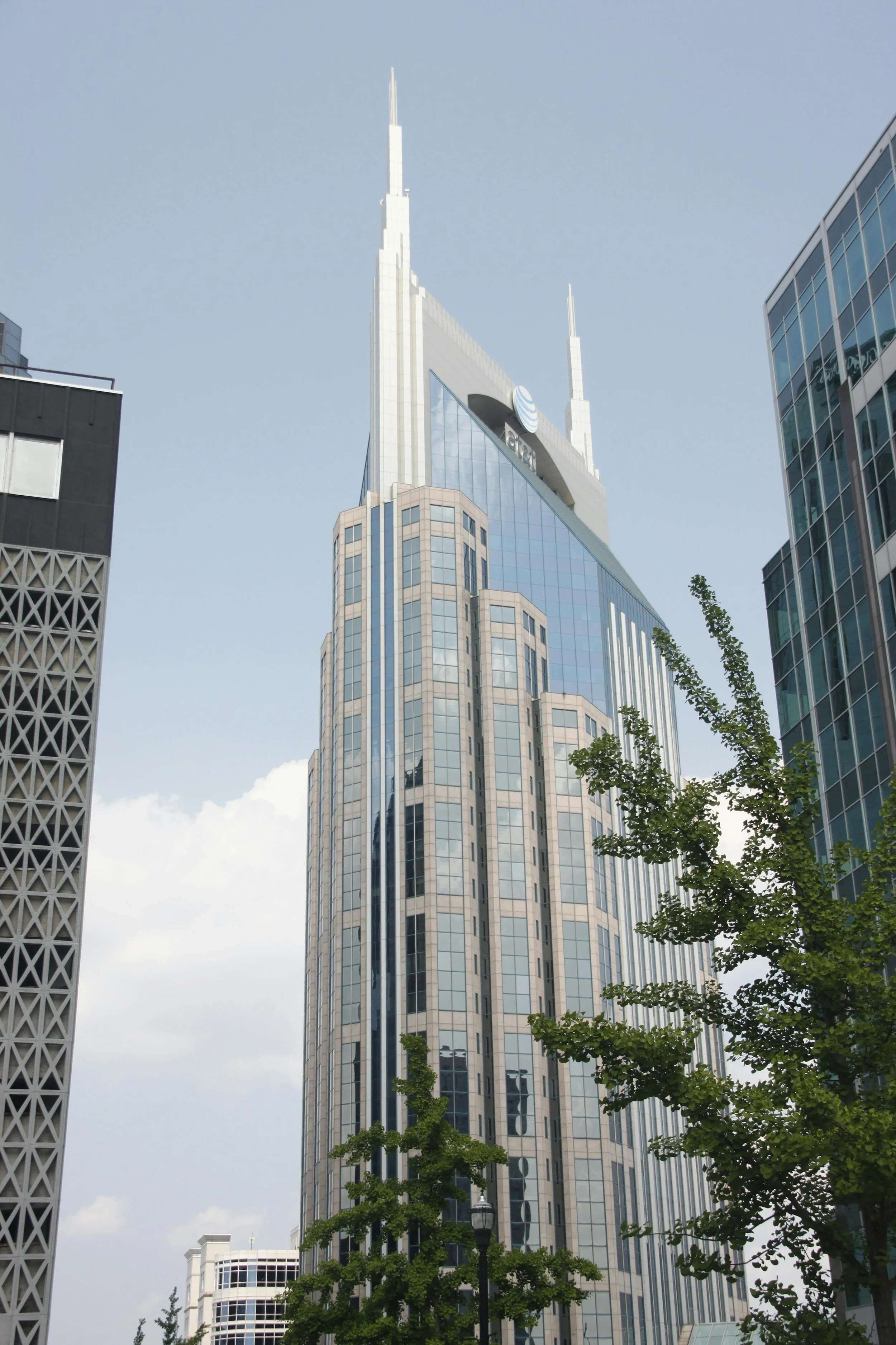 Skyline of a tall modern glass office building with AT&T logo at the top, surrounded by other skyscrapers and green trees in an urban setting.