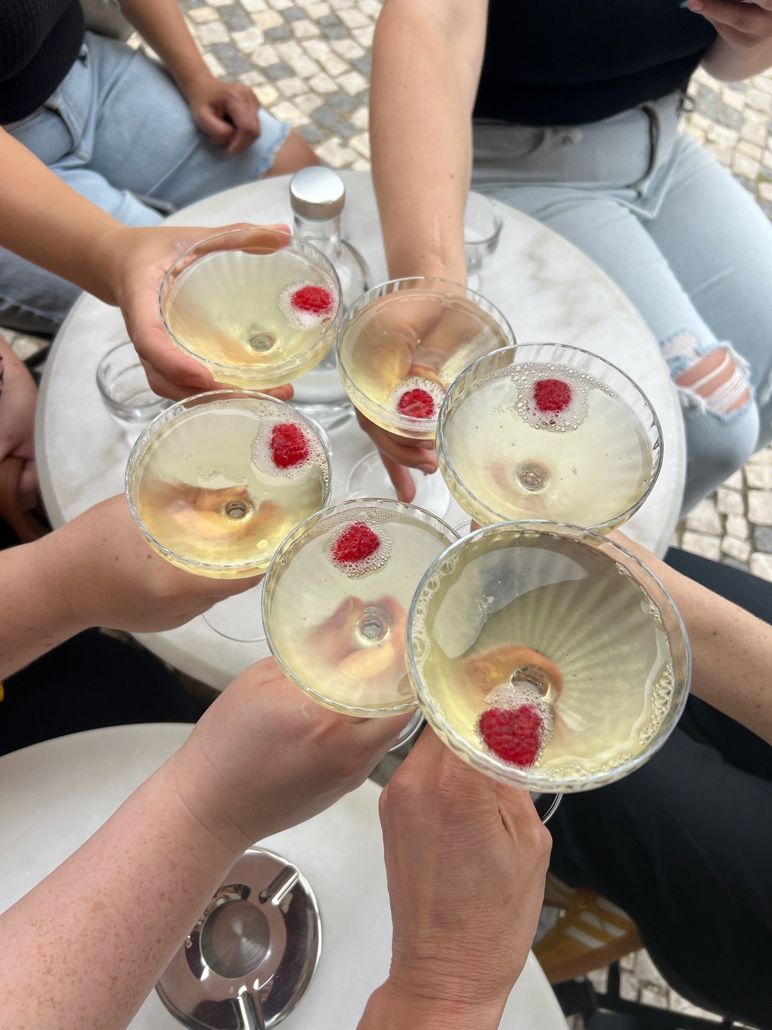 Several people clinking glasses of champagne with raspberries on top, seated around a white table.