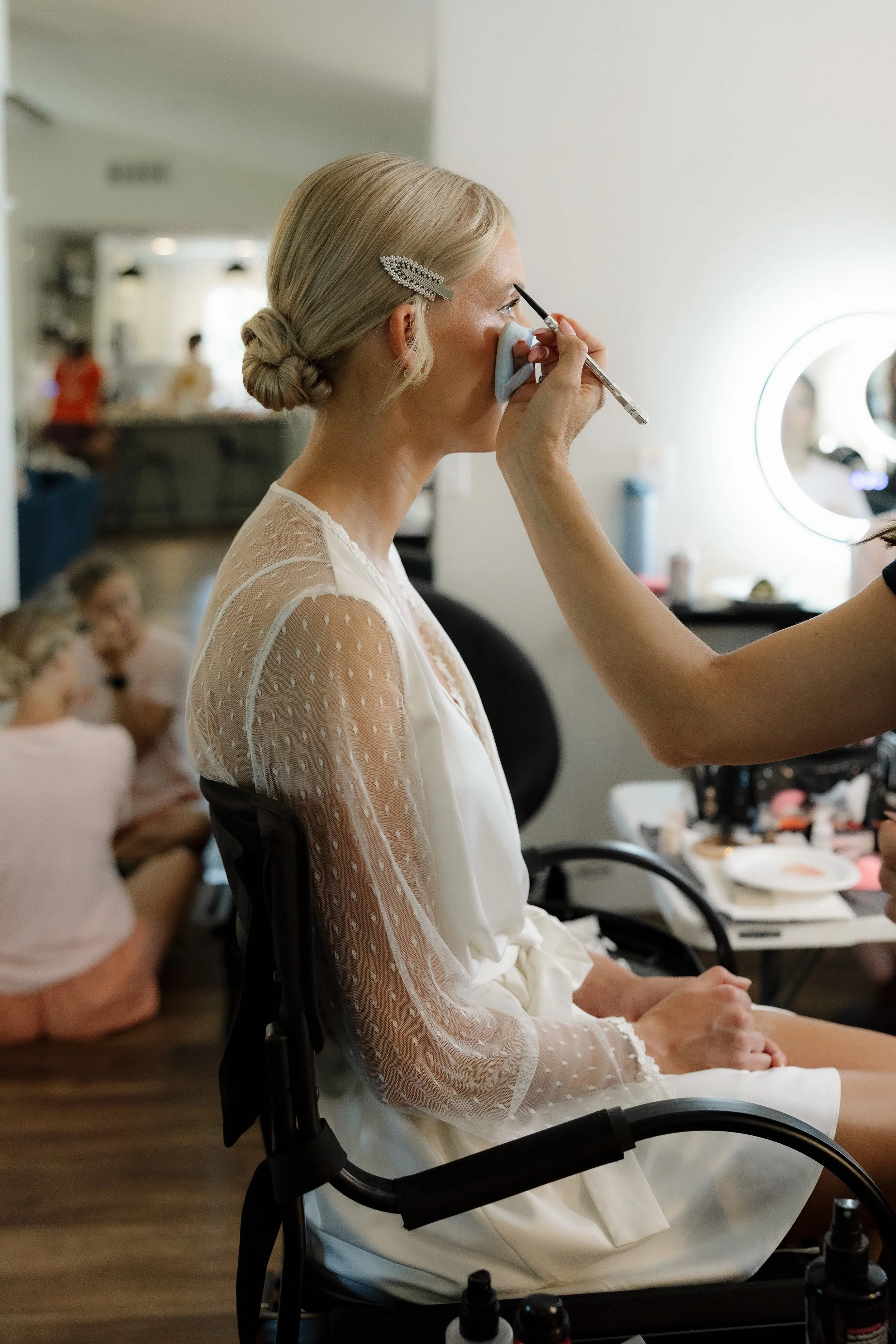 A woman with blonde hair styled in a bun, wearing a white, sheer, polka-dotted robe, is sitting in a chair while makeup artists applies makeup to her face in a room with other women preparing together.
