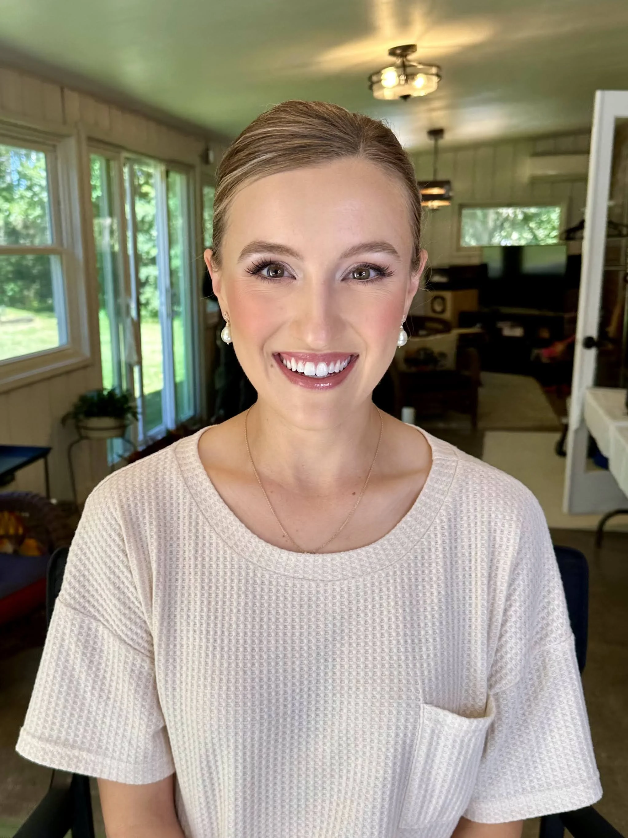Close-up photo of a young woman with brown hair and a bright smile, indoor setting with large windows and a modern design in the background.