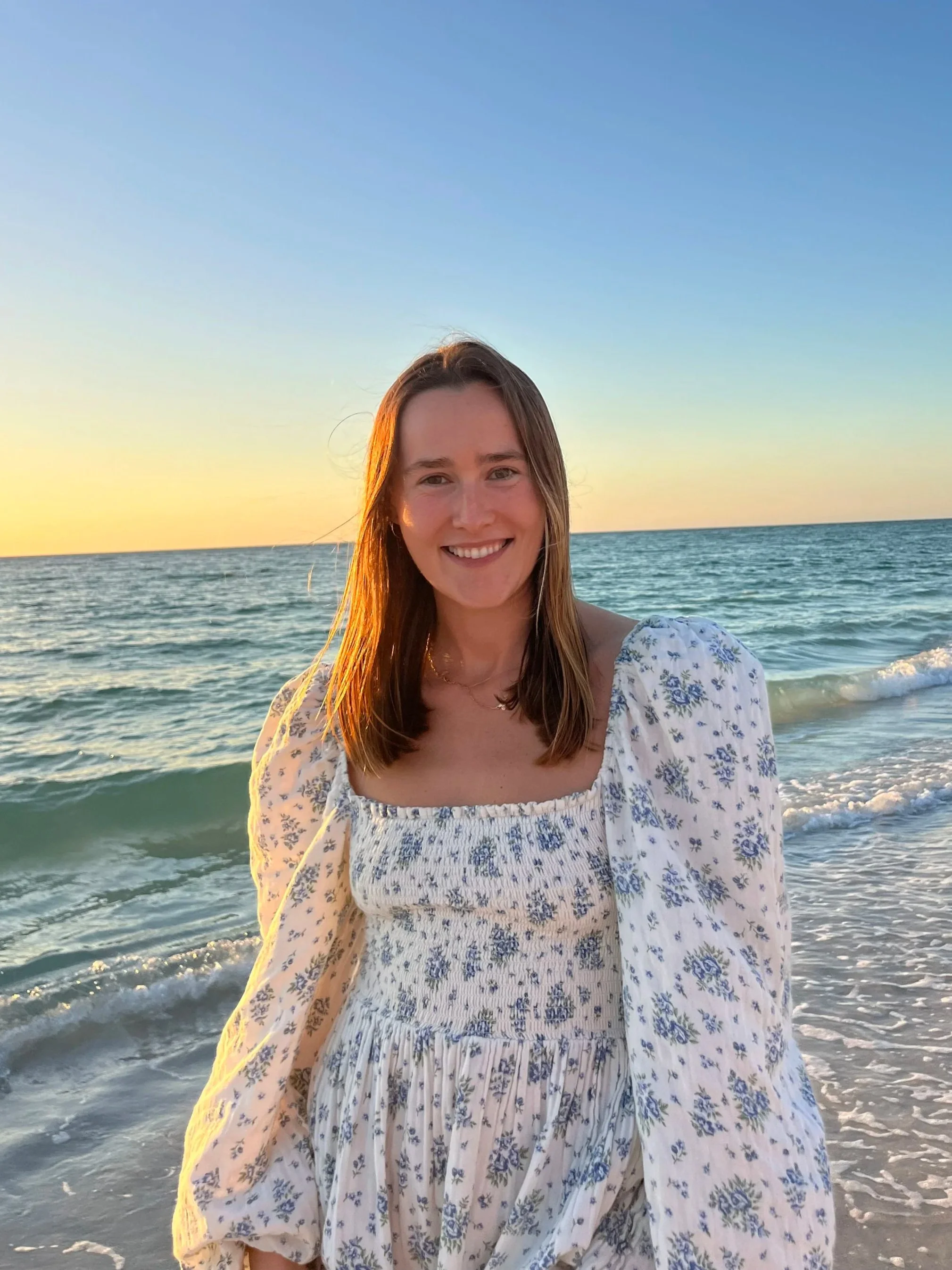 Young woman smiling at the beach during sunset, wearing a white dress with blue floral patterns.