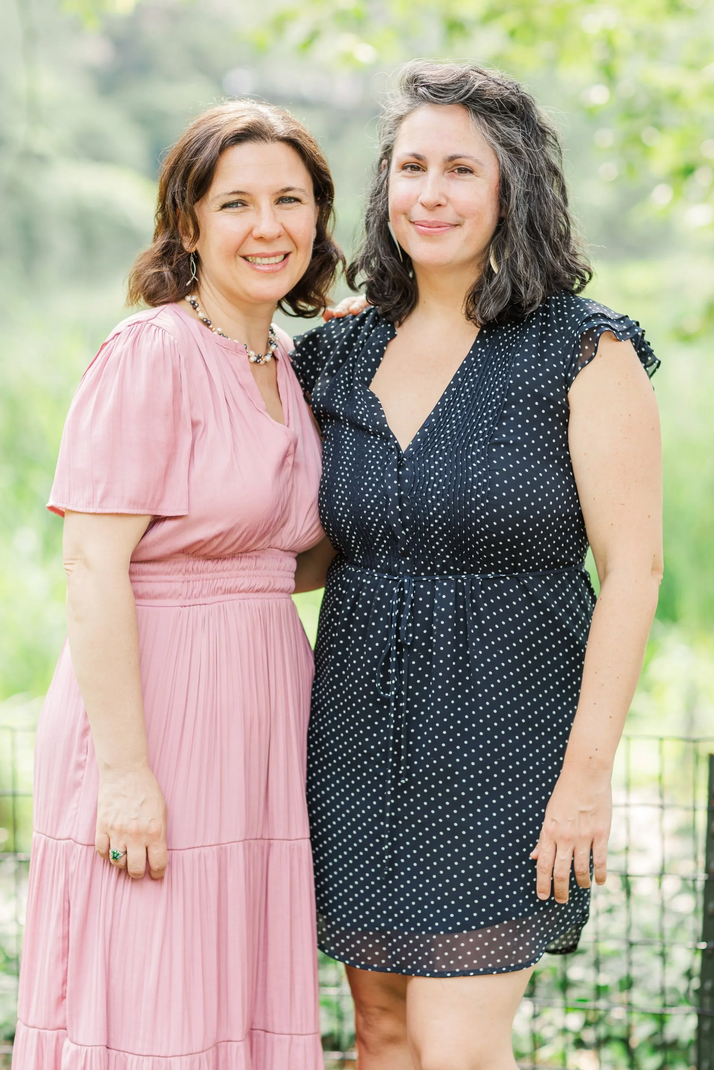 Two women standing outdoors on a sunny day with greenery in the background, smiling for the camera.