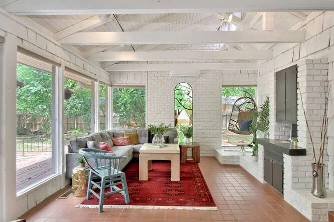 Bright living room with white brick walls, large windows, a gray sofa with colorful pillows, a blue armchair, a red area rug, a white coffee table, and hanging chair with cushions