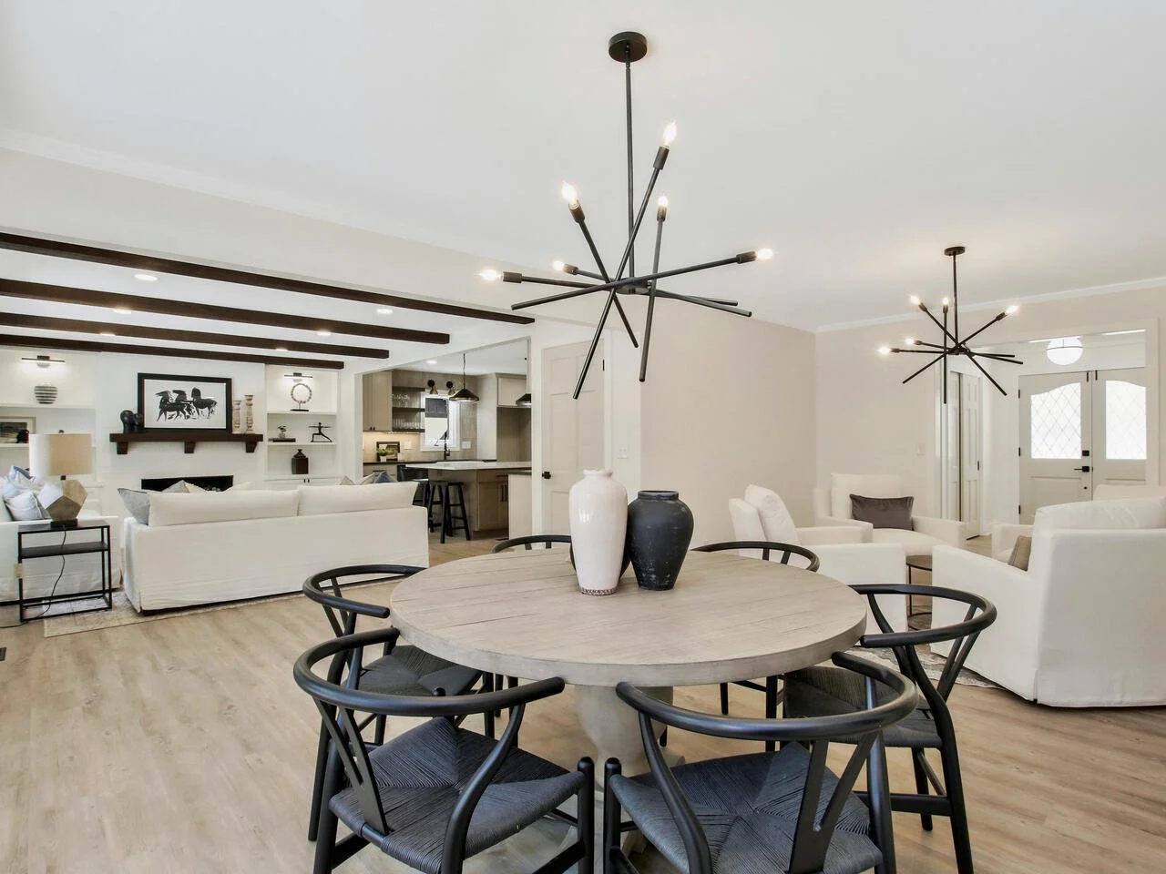 Open living room and dining area with modern black starburst chandeliers, white seating, and a round wooden dining table with vases.