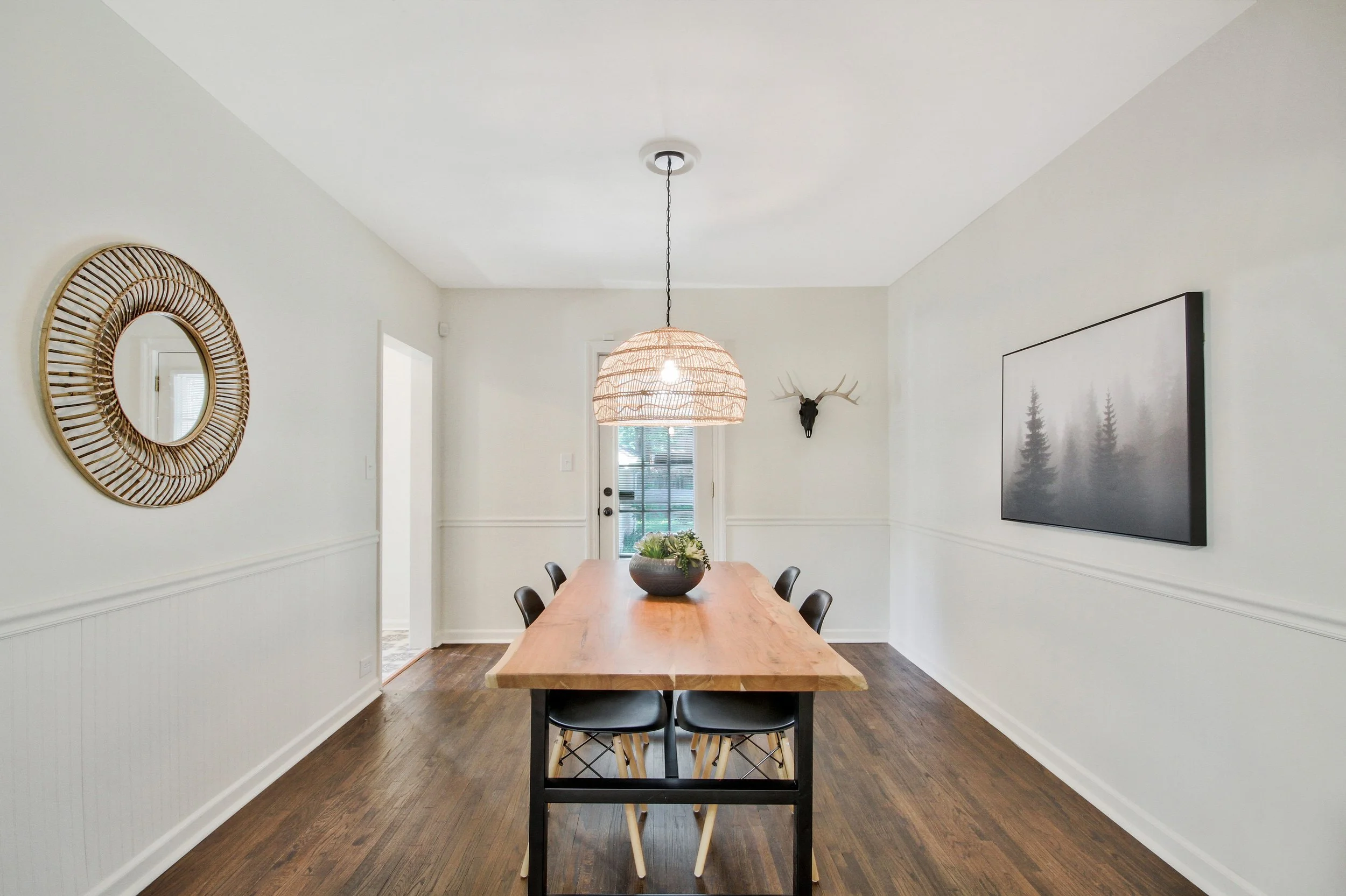 Dining room with a wooden table, six black chairs, a rattan pendant light, a wall-mounted TV displaying a forest scene, a mirror on the left wall, a mounted animal skull on the right wall, and a door leading outside at the back.