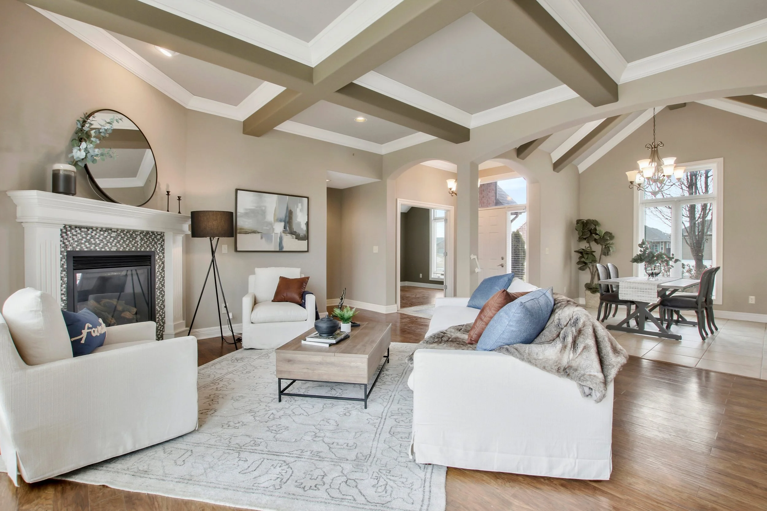 A spacious living room with a vaulted ceiling featuring exposed beams. It includes a fireplace with a large mirror and decorative items above, a white sofa with blue and brown pillows, an armchair, a wooden coffee table, and a patterned area rug. In the background, there's a dining area with a table, chairs, and a chandelier, along with large windows letting in natural light.