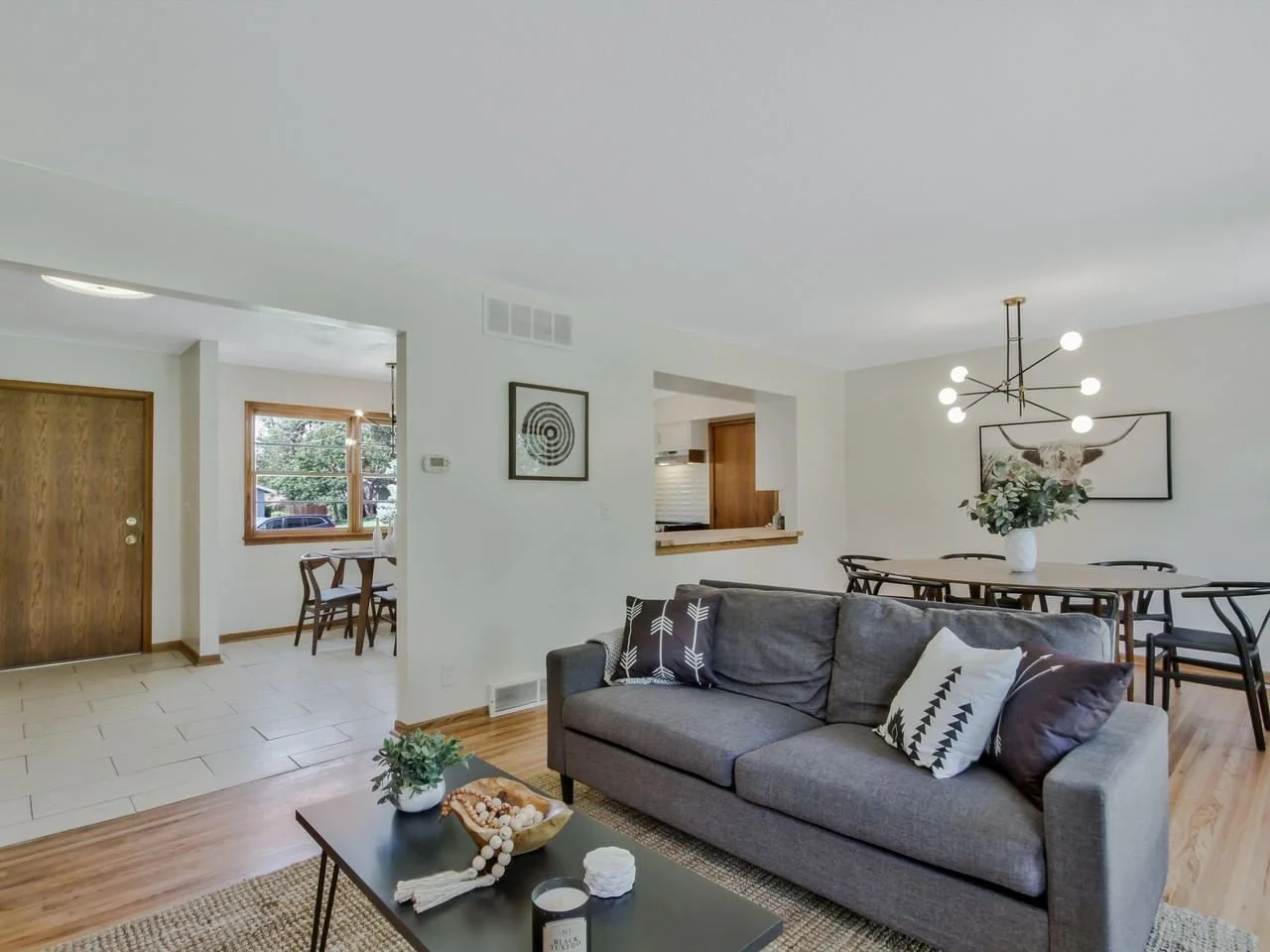 Living room with gray sofa, black and white pillows, wooden coffee table with a potted plant and decorative items, and a dining table with black chairs and a vase of flowers, with modern chandelier and framed artwork on walls.