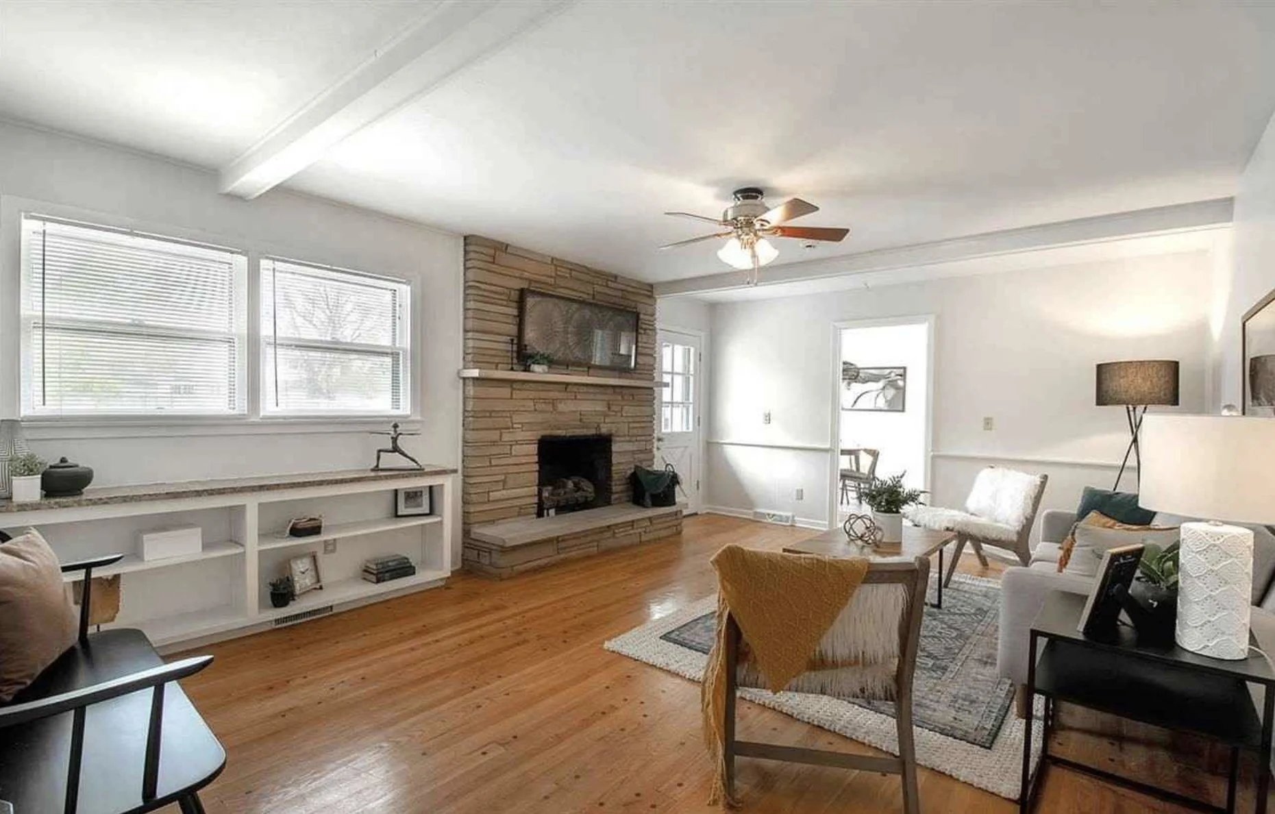 Living room with hardwood floors, a brick fireplace, large windows with blinds, white walls, and minimal decor. Furniture includes a white sofa, wooden chairs, a black side table, and a built-in white shelf. There is a ceiling fan with lights, a styled rug, and various decorative items.