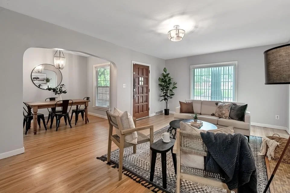 Living room with wooden floors, a beige sofa with throw pillows, a round coffee table, armchair, and large window with vertical blinds. There is a potted plant near the window and a small rug in front of the sofa. Adjacent dining area with a wooden table, black chairs, and a round mirror on the wall.