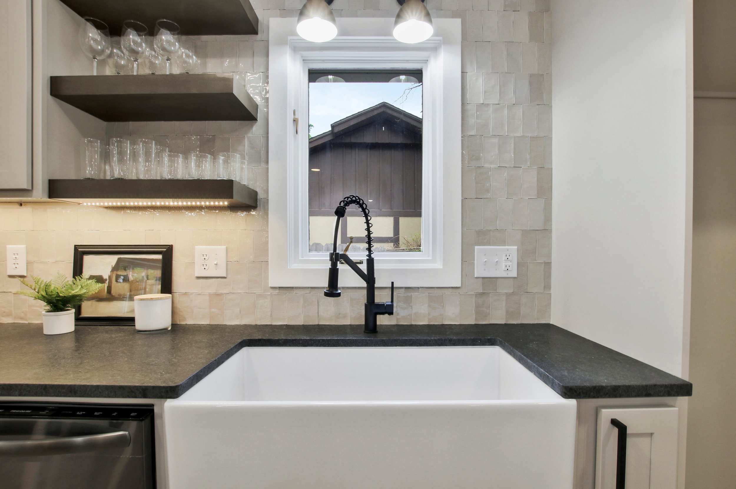 Modern kitchen sink with black faucet, white farmhouse-style basin, granite countertop, window showing house roof outside, open shelves with glasses, decorative plant and picture frame, beige tiled backsplash, and white cabinet.