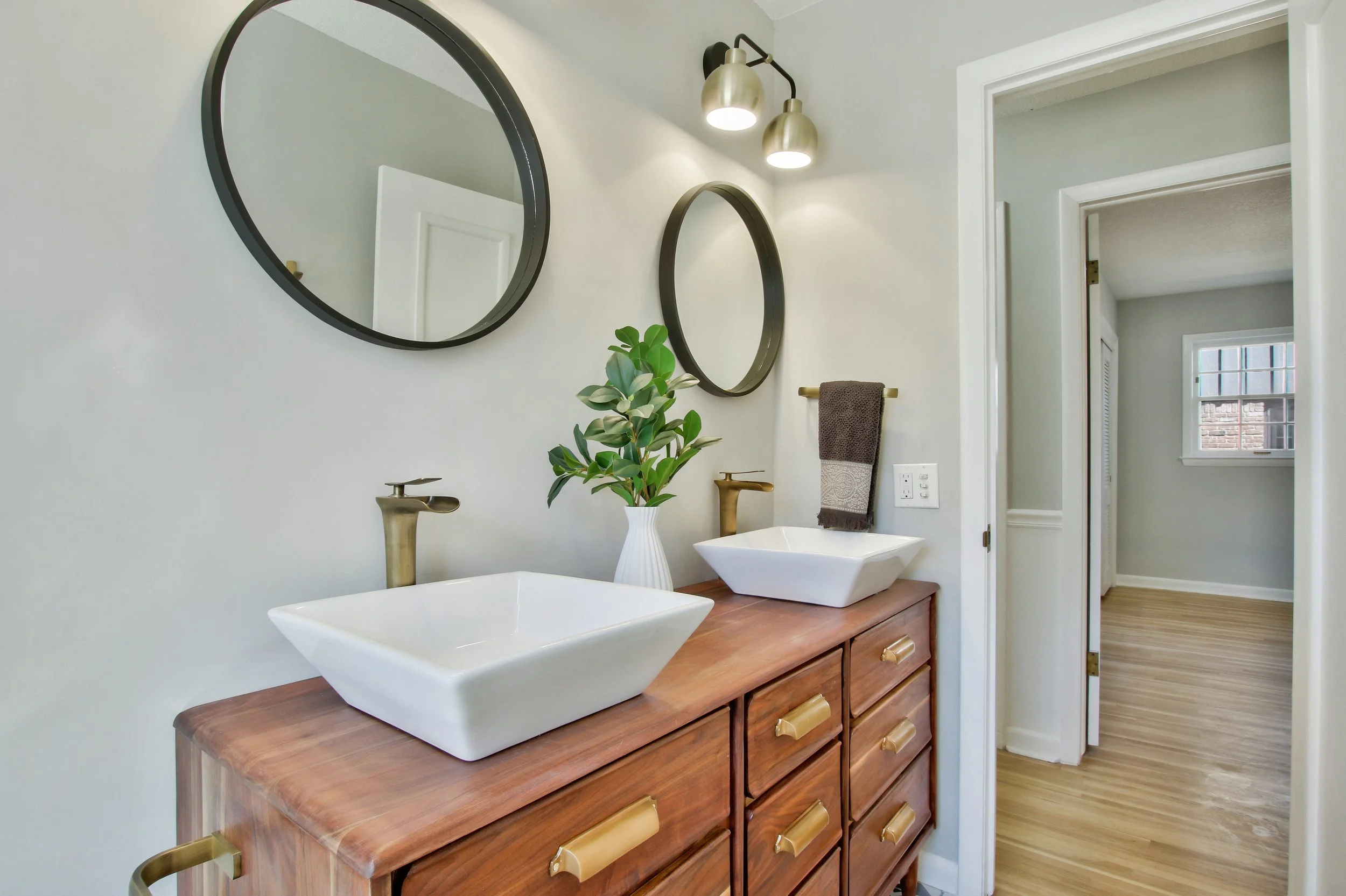 Bathroom with double vessel sinks on a wooden vanity, two round mirrors on the wall, two gold faucets, a vase with green plants, and a dark towel on a towel rack.