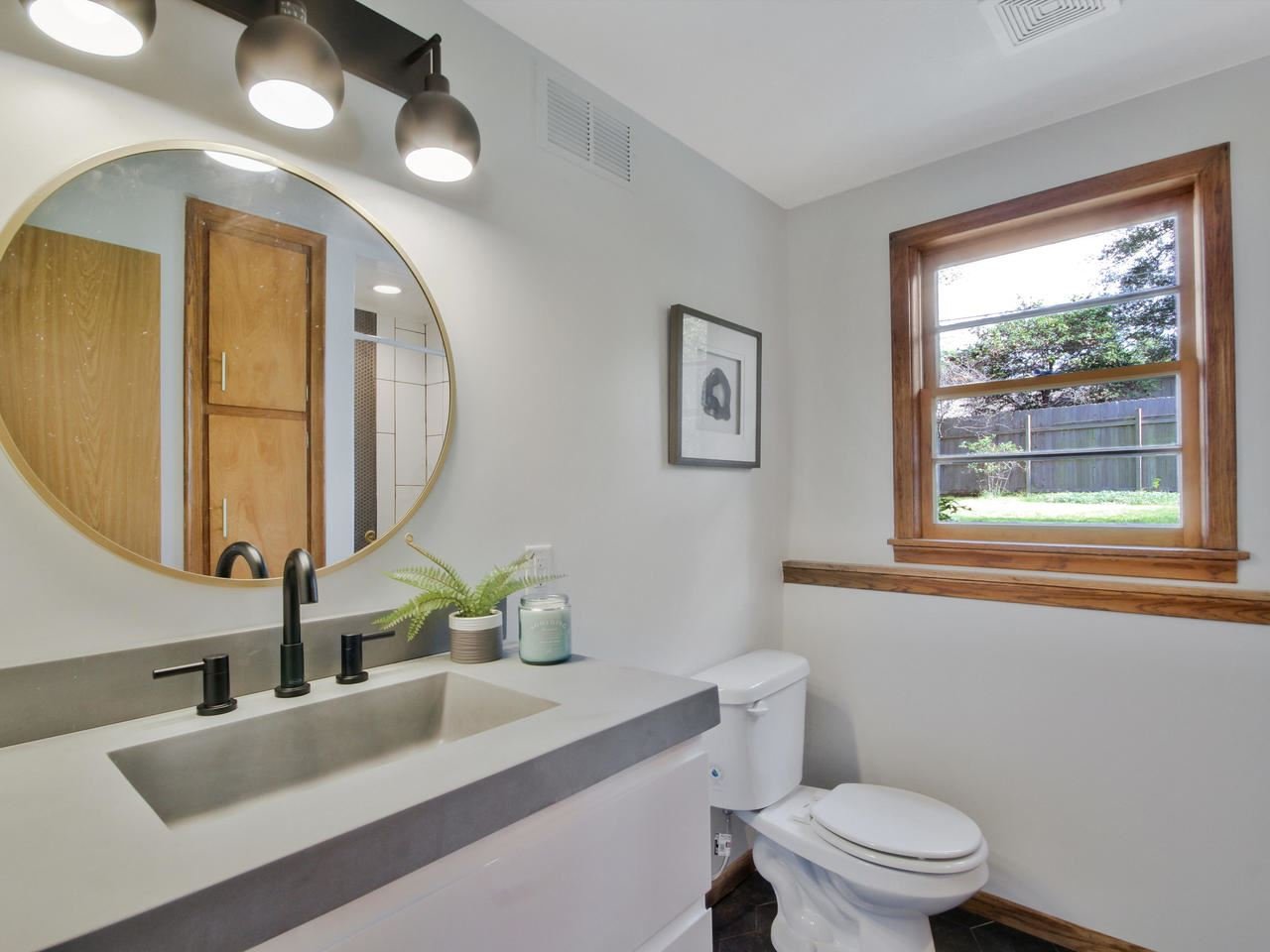 A bathroom with white walls, a round mirror above a concrete countertop with a black faucet, decorated with a small plants and candles. To the right is a toilet, and a window with wooden trim showing a backyard with green grass and trees.