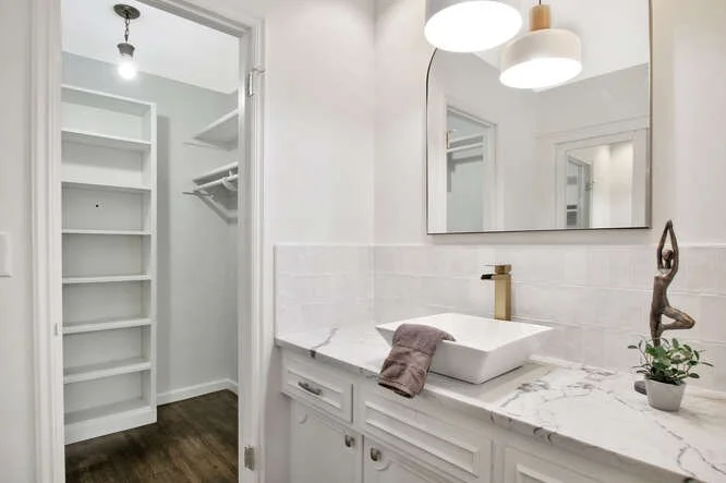 Bathroom vanity area with a marble countertop, white vessel sink, gold faucet, decorative sculpture, plant, and mirror; walk-in closet with built-in shelves and a hanging rod