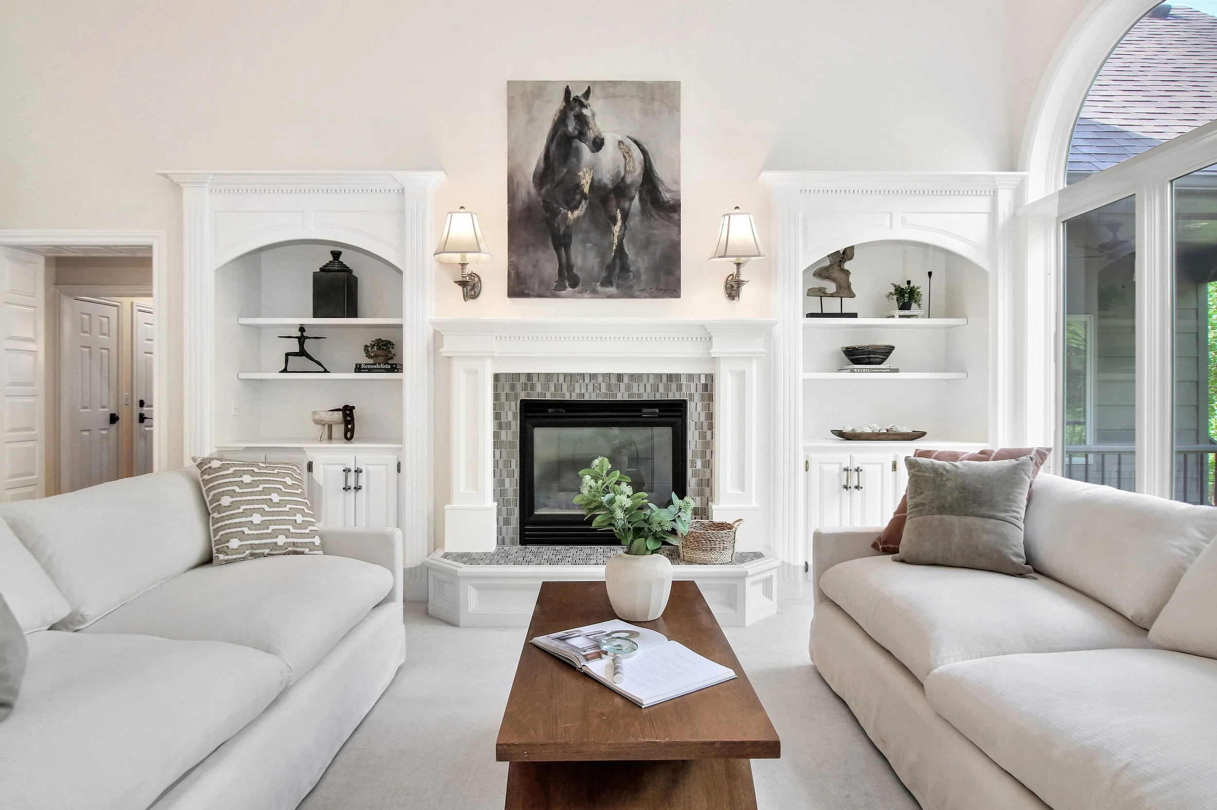 Living room with white sofas, a fireplace with a painting of a horse above, built-in shelves on either side, and a wooden coffee table with a vase of greenery and magazines.