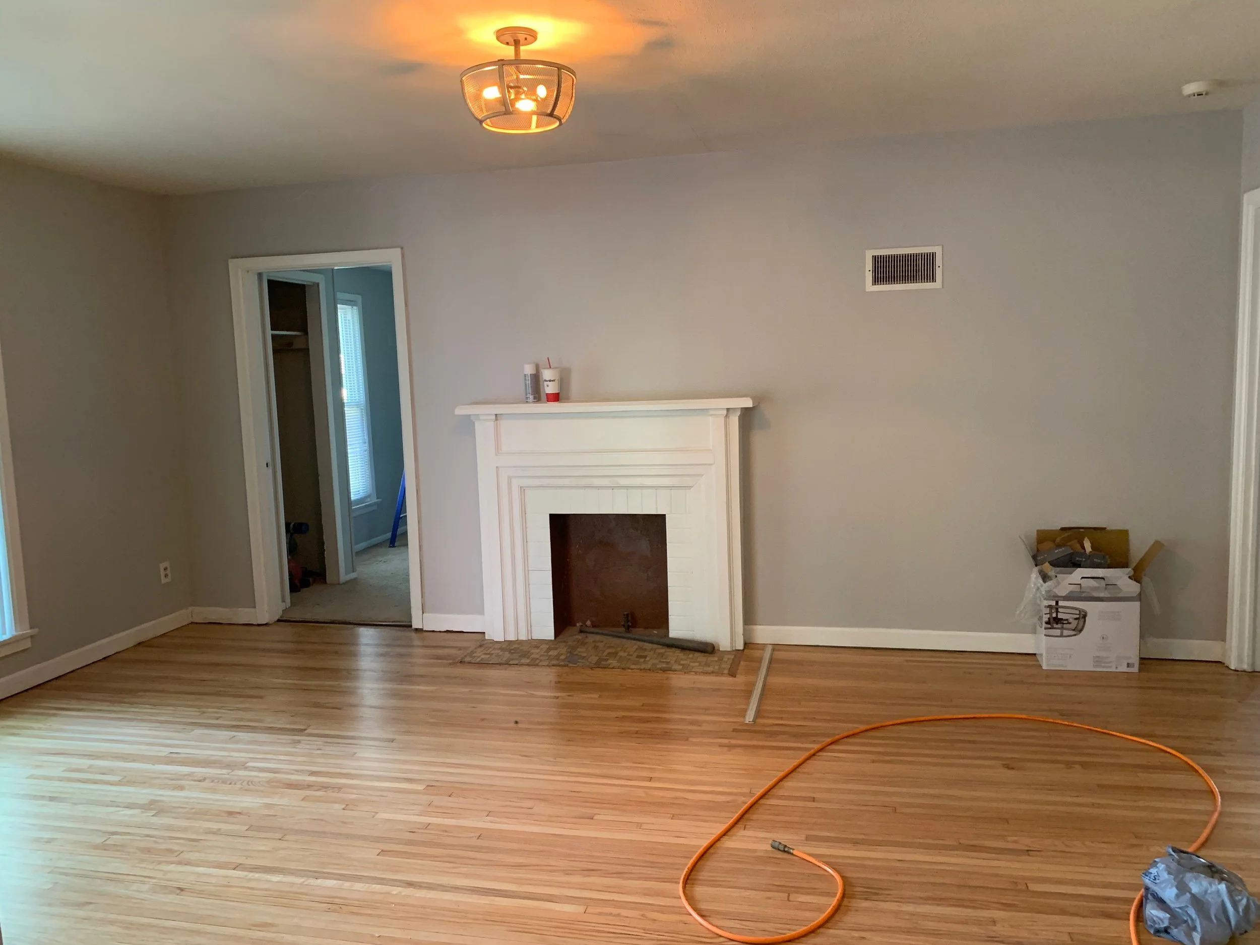 Empty living room with light wood floor, gray walls, and a white fireplace with an open space for a fireplace insert, with items on the mantle. An orange extension cord is on the floor to the right, and a box with tools is in the corner. An open door to a small room or closet is on the left, with a window in that room.