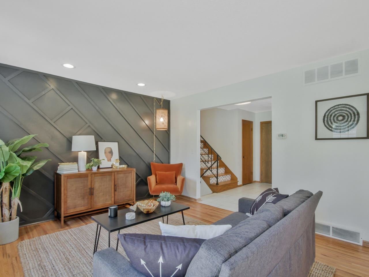 Living room with a gray sofa, orange armchair, black coffee table, and a wooden sideboard against a black accent wall with diagonal paneling. There is a large plant on the left, a framed picture, a lamp, and some decor. An open staircase with wooden steps and metal railing leads upstairs, and the room has hardwood floors, white walls, and minimalist artwork.
