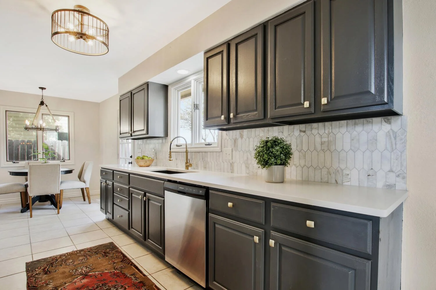 Modern kitchen with dark gray cabinets, white marble backsplash, white countertop, and stainless steel dishwasher. There is a small potted plant on the counter and a bowl of green apples near a window.