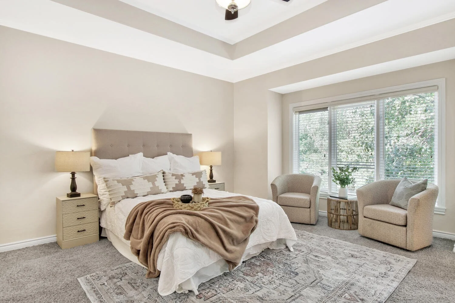 A cozy bedroom with a large bed featuring a padded headboard, white bedding, and a beige throw blanket. Two bedside tables with matching lamps flank the bed. There are two light-colored armchairs by a large window with blinds, a potted plant on a side table, and a patterned area rug on the carpeted floor.