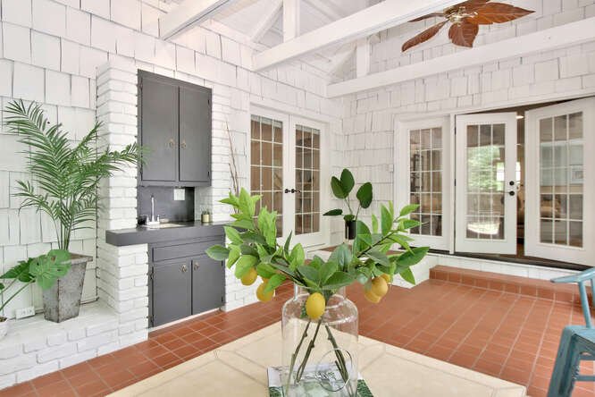 Bright sunlit porch with white brick walls, a gray cabinet, a potted plant, glass doors, and a glass vase with a lemon and leafy branches on a table.