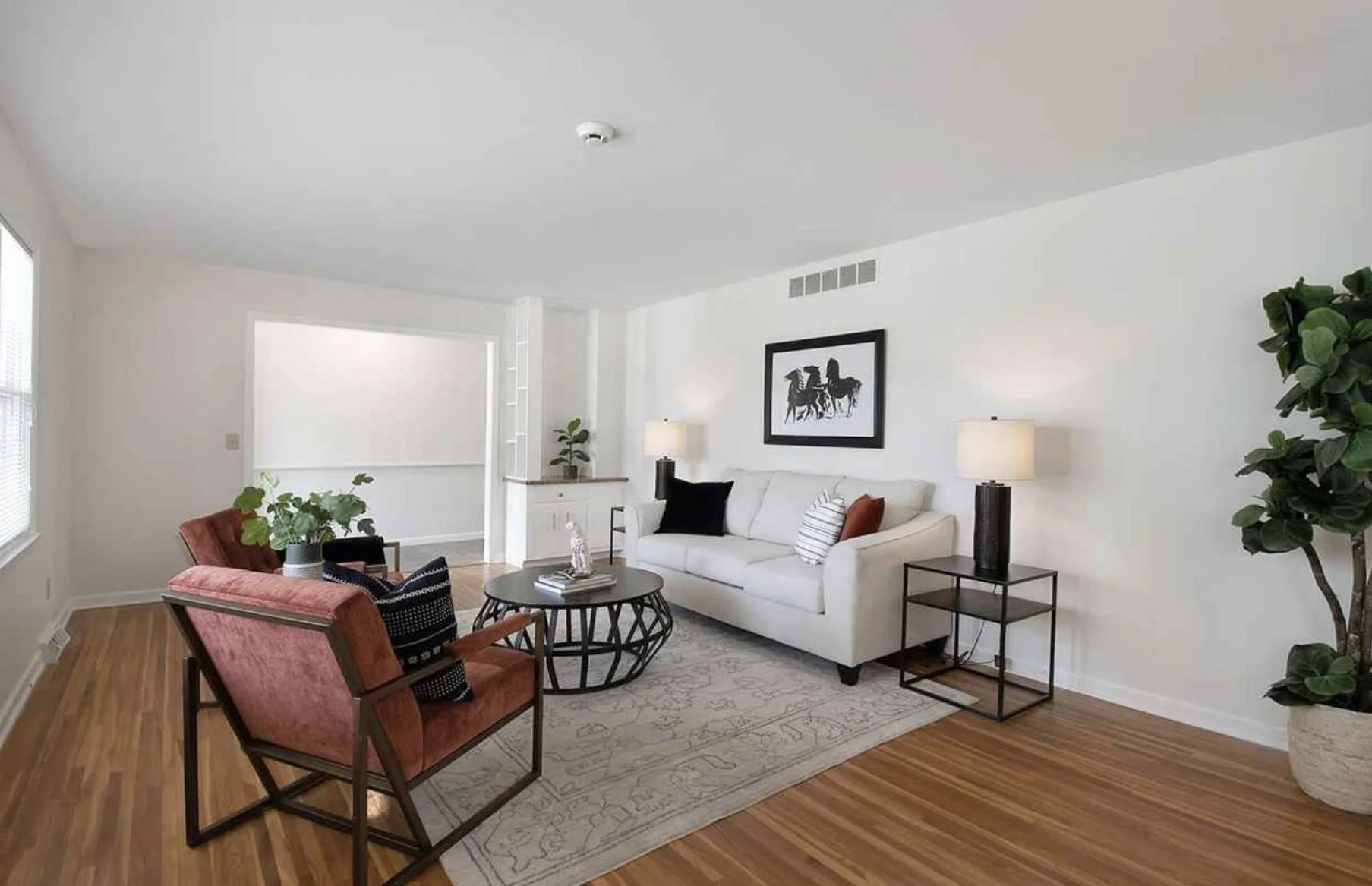 Living room with white walls, hardwood floors, a white sofa, and two side tables with lamps. There are two armchairs, one upholstered in coral fabric and the other in green, with pillows. A round black coffee table, a small white cabinet, and a large potted plant are also visible.