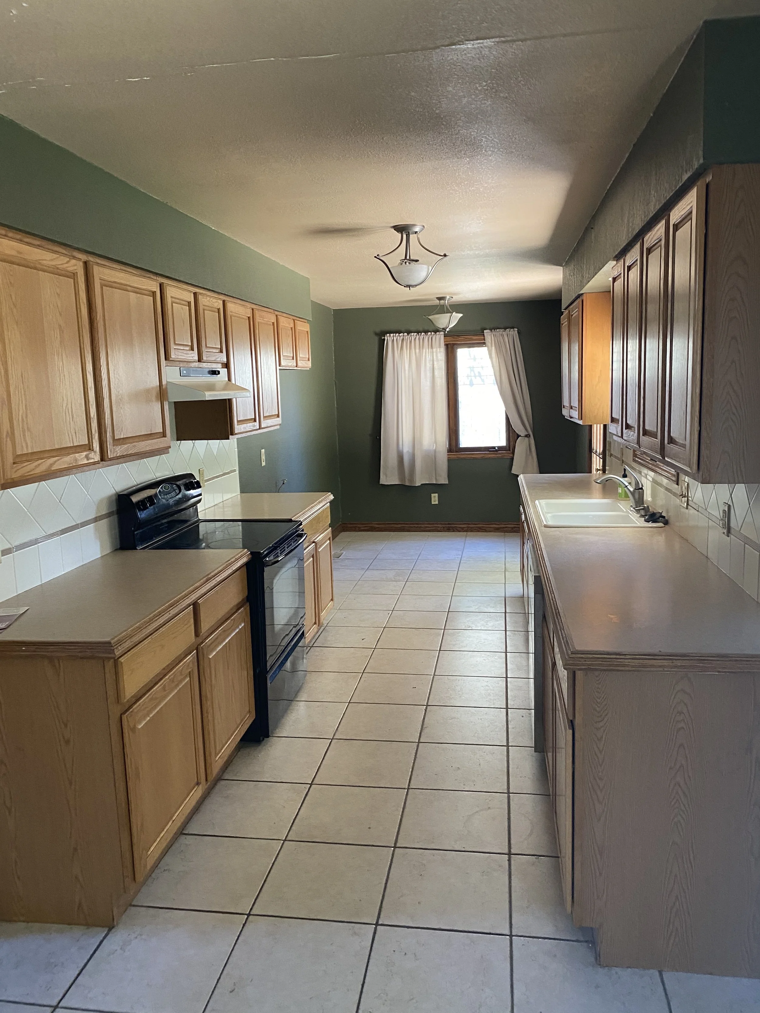 Empty kitchen with wooden cabinets, beige countertops, a black stove, a white sink, a window with white curtains, and tiled floor. Two ceiling lights.