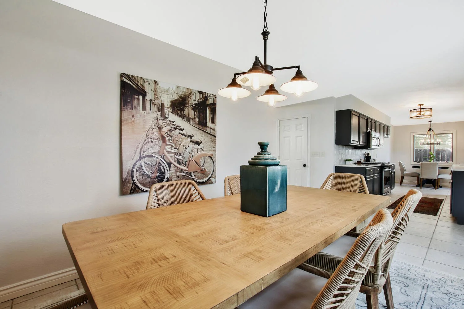 A dining room with a large wooden table, rattan chairs, a picture of bicycles on the wall, a hanging light fixture, and a view into the kitchen and adjacent dining area with a window and additional lighting.
