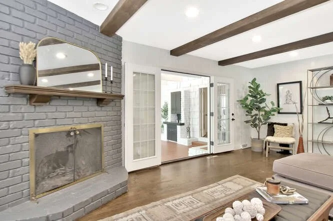 Living room with gray brick fireplace, wooden mantel, large mirror, glass doors leading outside, beige sofa, patterned rug, bookshelf with decorative items, and a large potted plant.