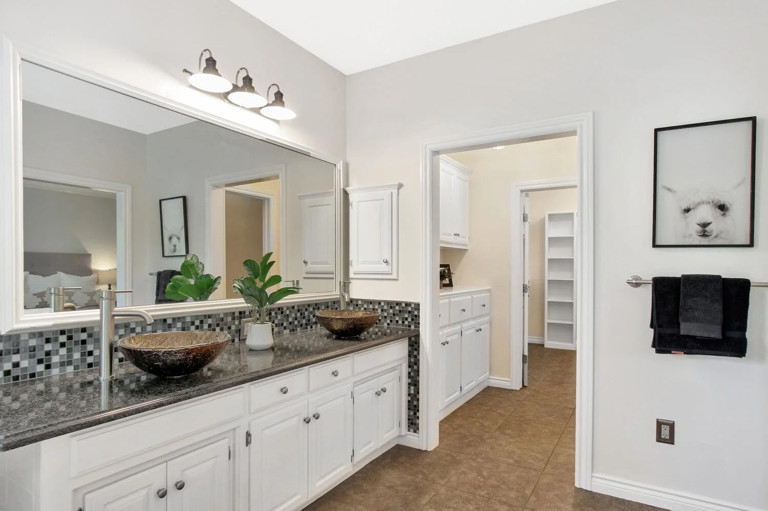 Modern bathroom with white cabinets, granite countertop, double vessel sinks, large mirror, black and gray backsplash, wall-mounted light fixture, black towels, framed llama picture, and a side door leading to a closet with shelves.