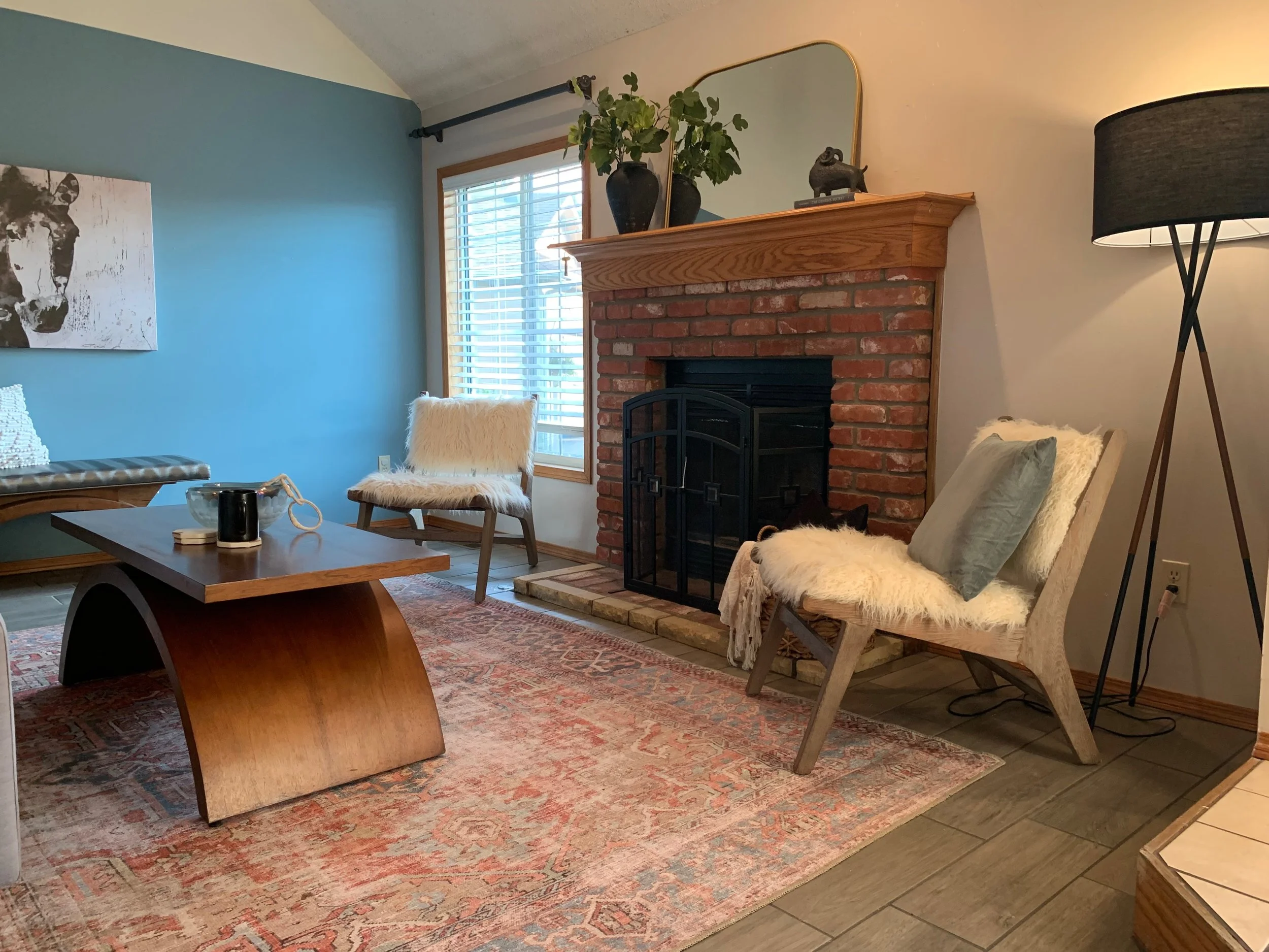Living room with a brick fireplace, two cream-colored chairs with fur throws and pillows, a wooden coffee table, a blue accent wall with a cow painting, a window with blinds, a tall floor lamp, and decorative items on the mantel.