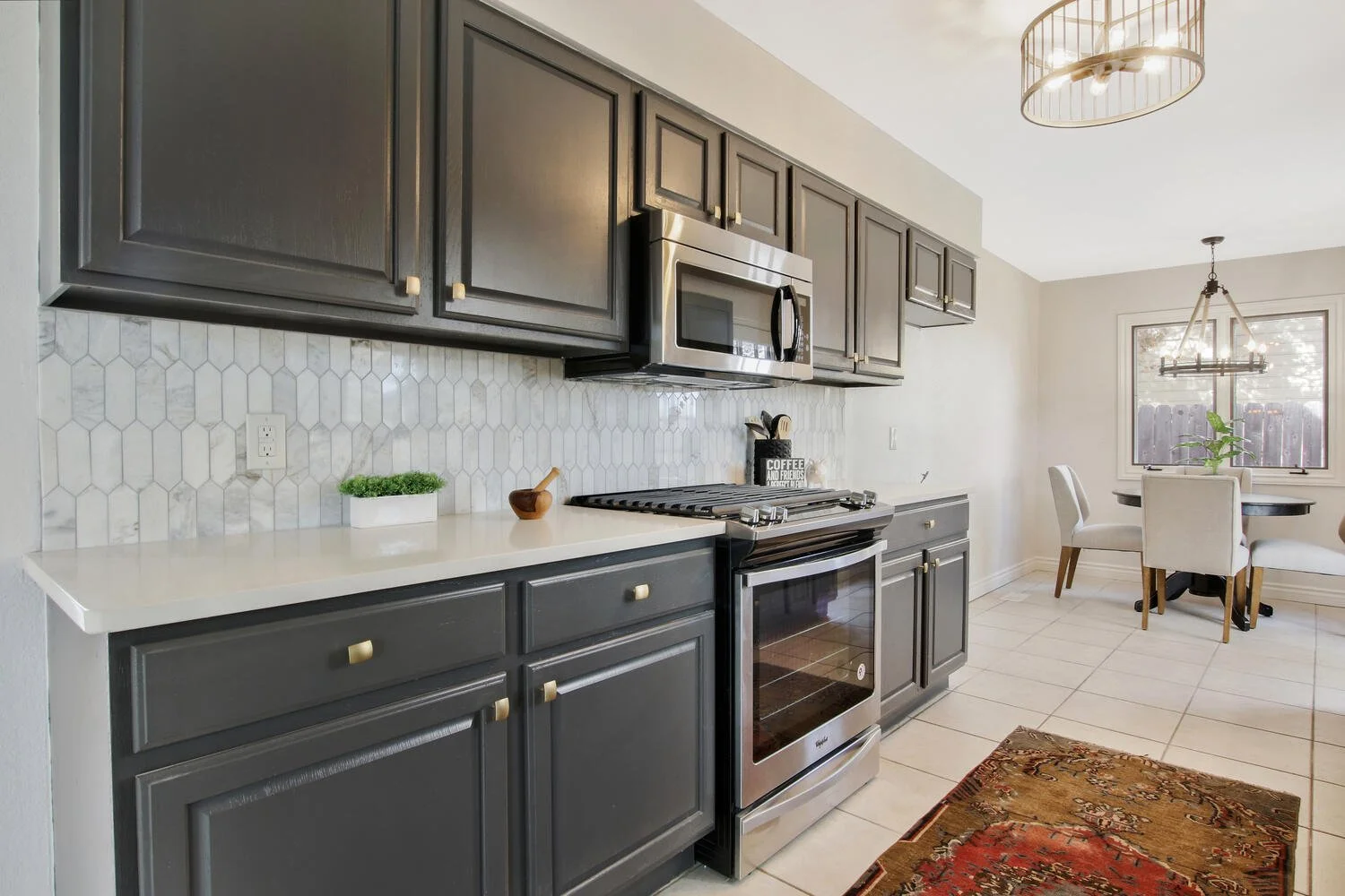 Modern kitchen with dark gray cabinets, white countertop, stainless steel microwave, oven, gas stove, and a small dining area with four white chairs and a round table near a window.