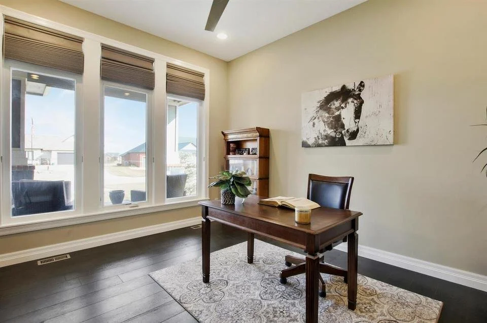 Home office with large windows, a wooden desk with a book, a coffee, a chair, a small bookshelf, a plant, and a horse canvas on the wall.