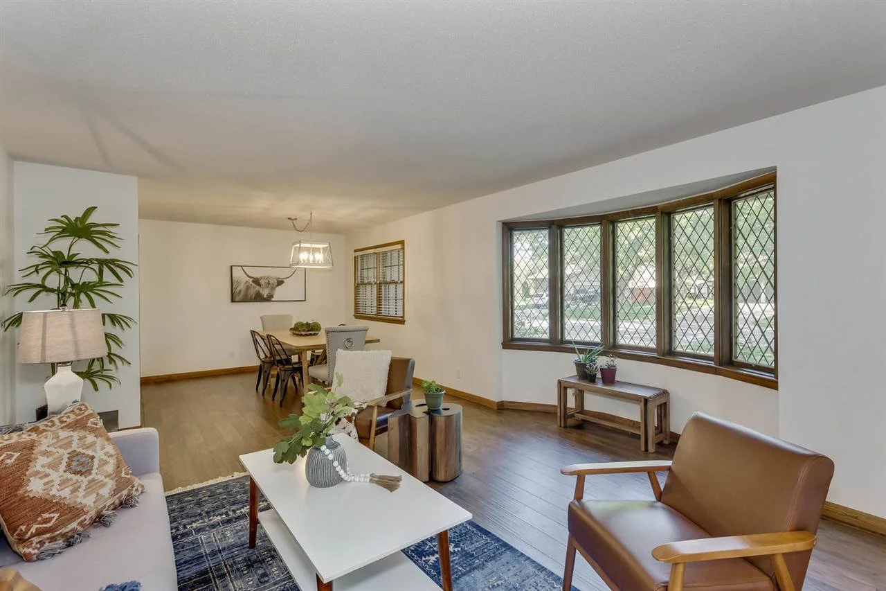 Living room with wooden flooring, a large window with diamond grid pattern, a white sofa with patterned pillow, a armchair, and a dining table with chairs. Decor includes plants, a ceiling light, and wall art of a cow.