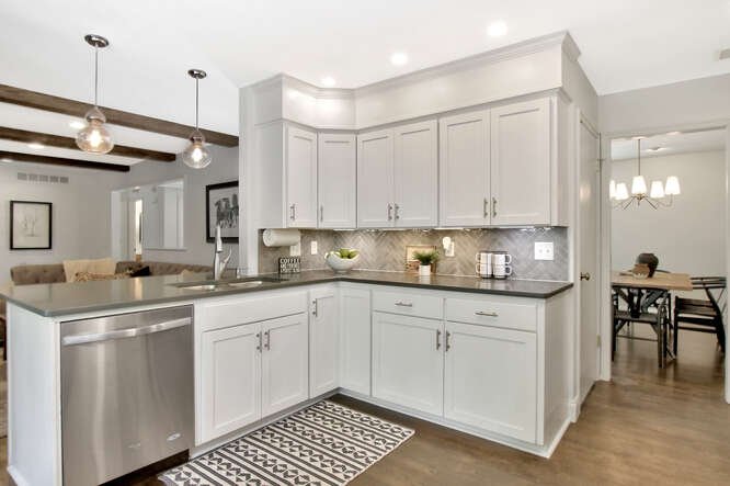 Modern kitchen with white cabinets, gray countertops, stainless steel dishwasher, and pendant lighting, adjacent to dining area.