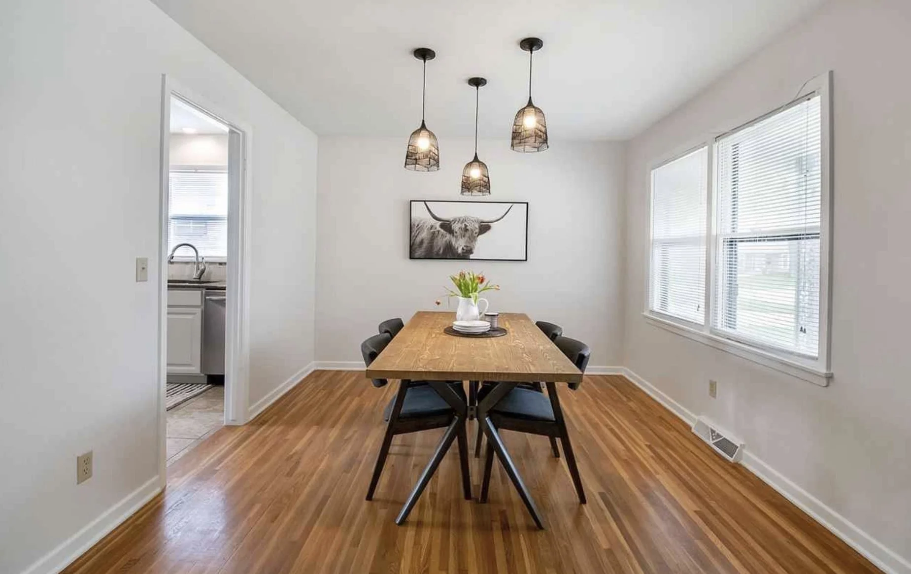 A dining room with a wooden table, six black chairs, a white vase with flowers, a framed black and white highland cow photo, large windows with blinds, three hanging pendant lights, and hardwood floors.