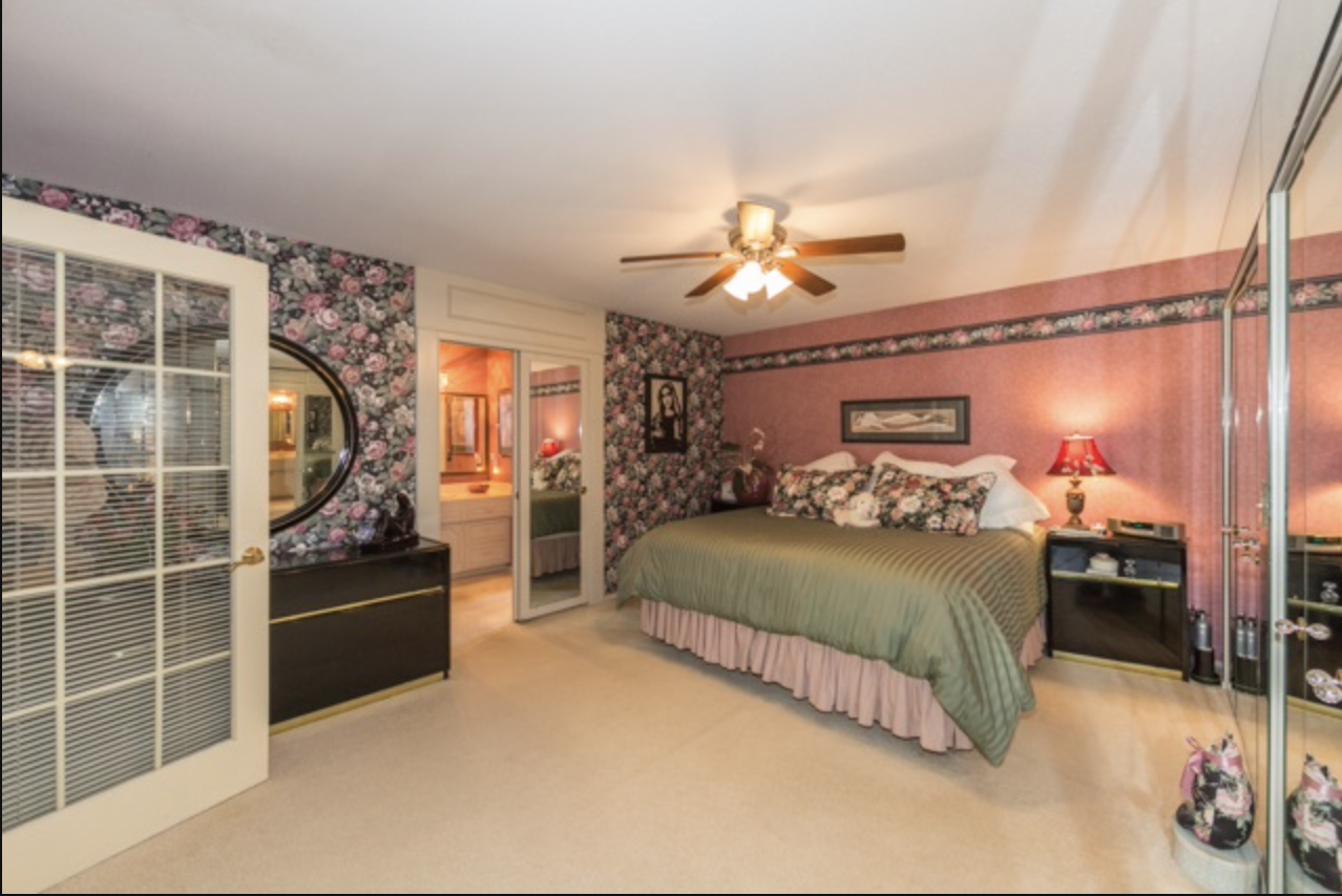 A bedroom with floral wallpaper, a bed with a green bedspread, pink ruffled bed skirt, and floral pillows, a ceiling fan, a mirrored closet door, two black nightstands, a red lamp, and a framed picture above the bed.