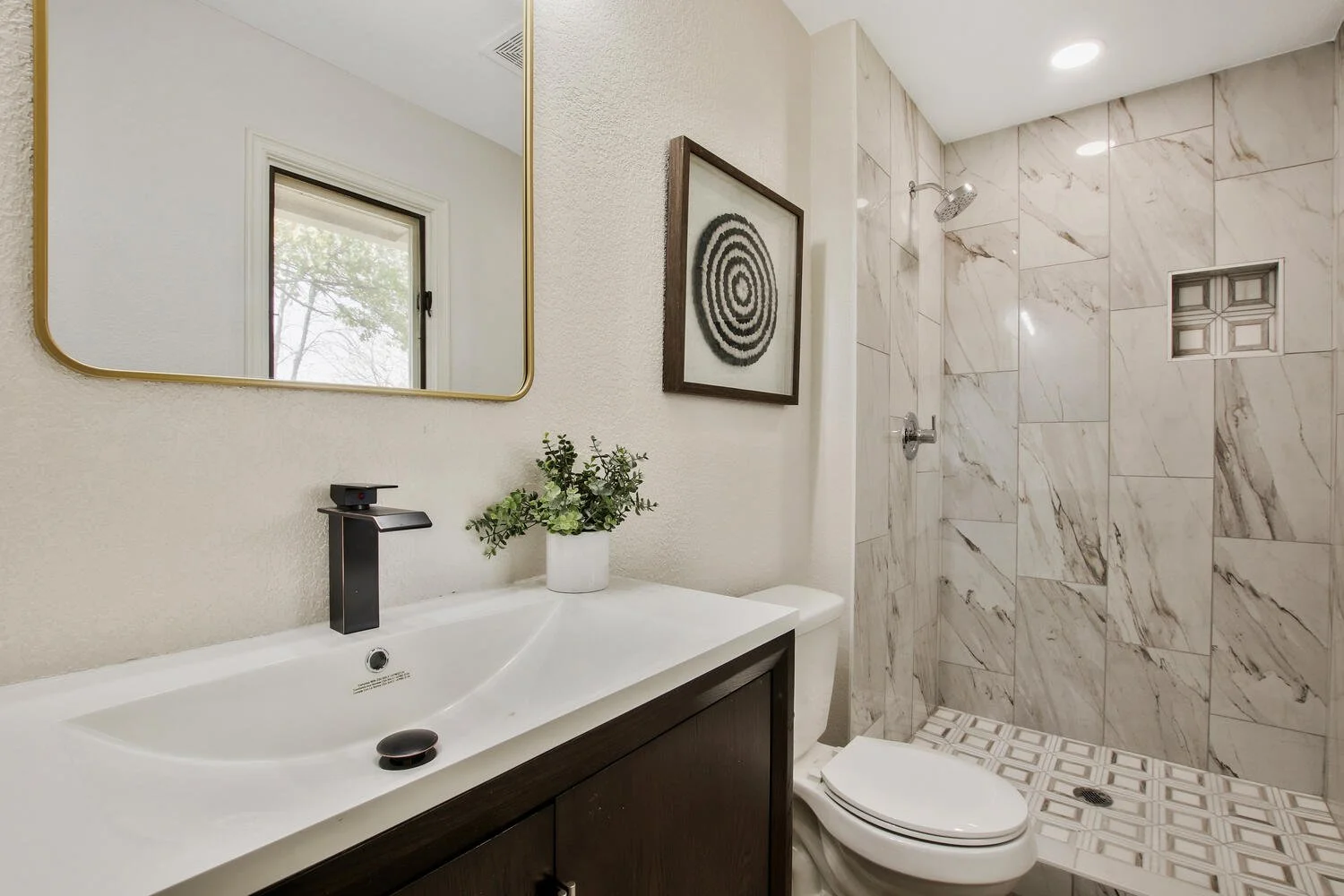 Modern bathroom with a white vanity, black faucet, potted plant, framed black-and-white artwork, toilet, and a walk-in shower with marble tiles and a small shower niche.