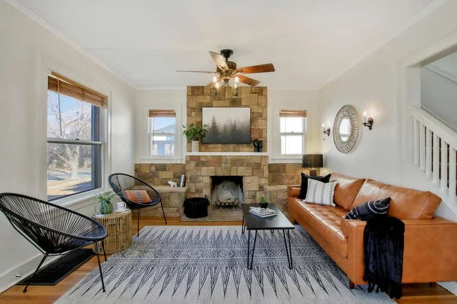 Living room with a stone fireplace, a mounted TV, two black chairs, a brown leather sofa, a black coffee table, and a patterned rug.