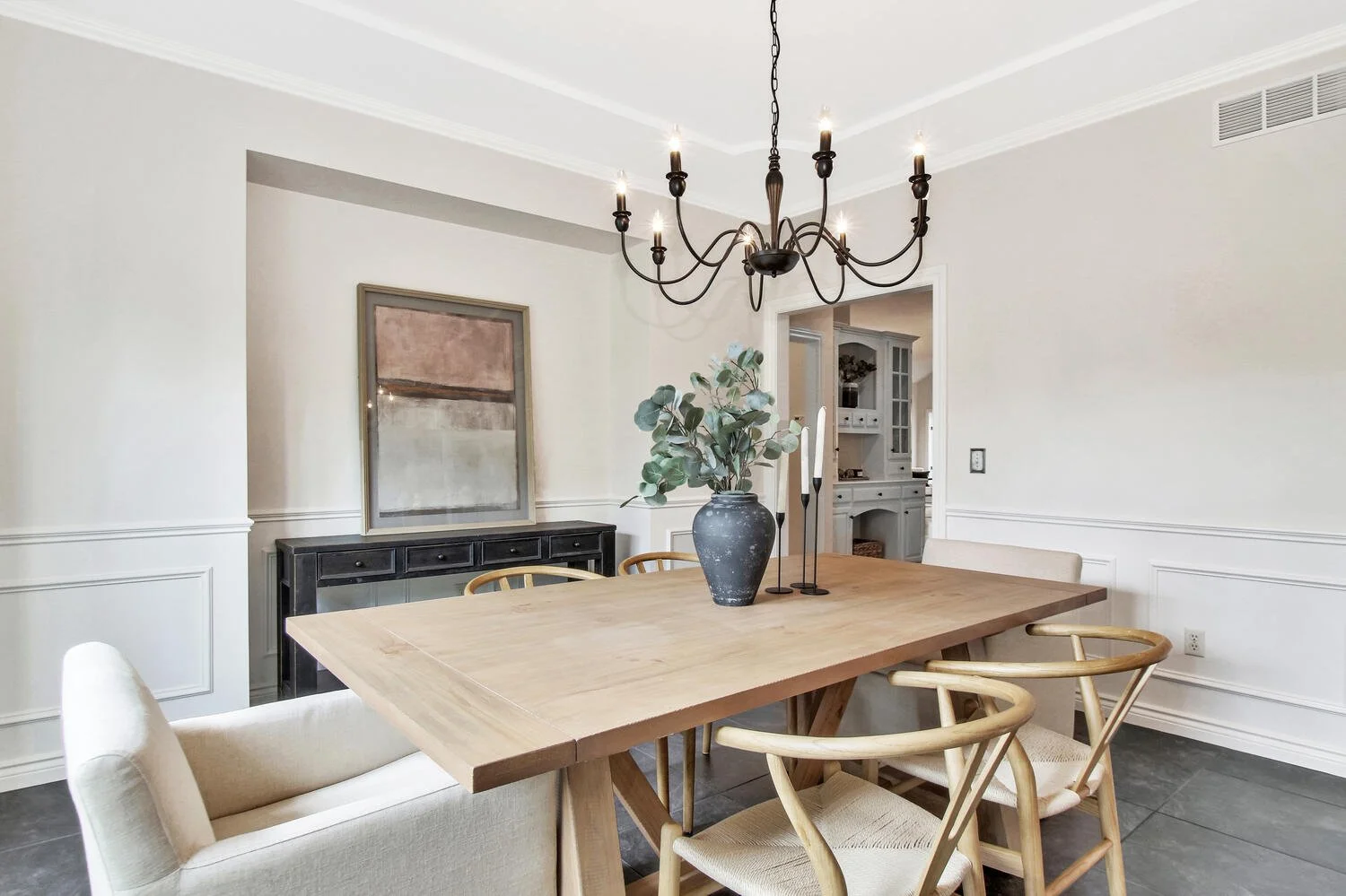 A dining room with a wooden table, beige chairs, a black chandelier, a large vase with green leaves, and white walls with decorative molding.