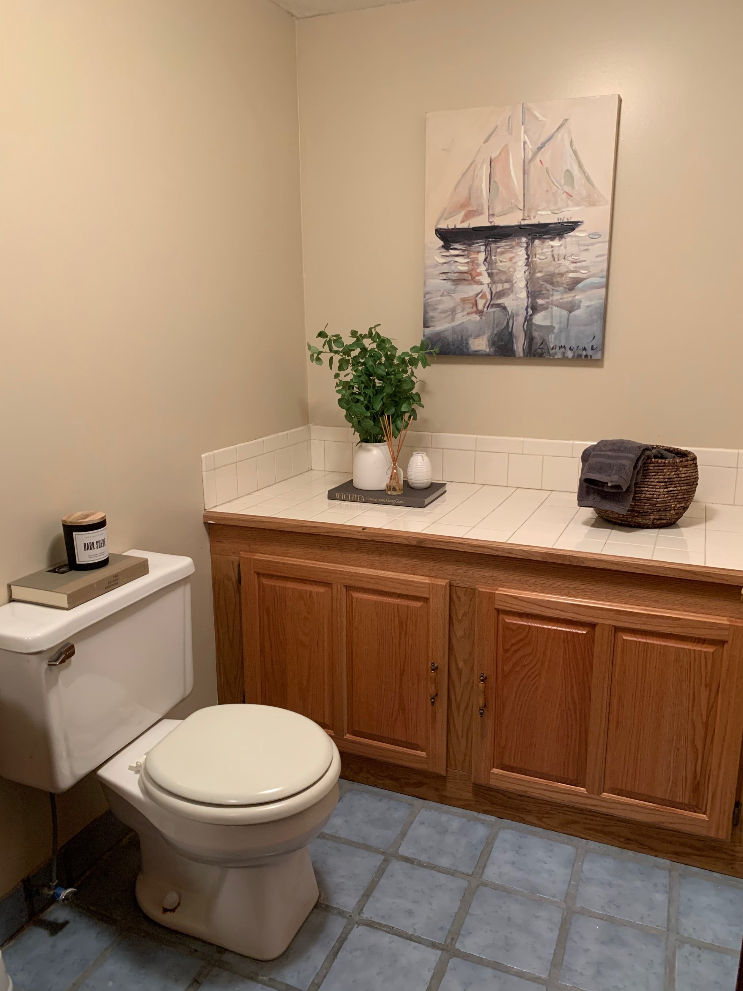 A bathroom with a white toilet and a wooden cabinet with a tiled countertop. Decor includes a potted plant, candles, a book, a basket with folded towels, and a painting of a sailboat on the wall.