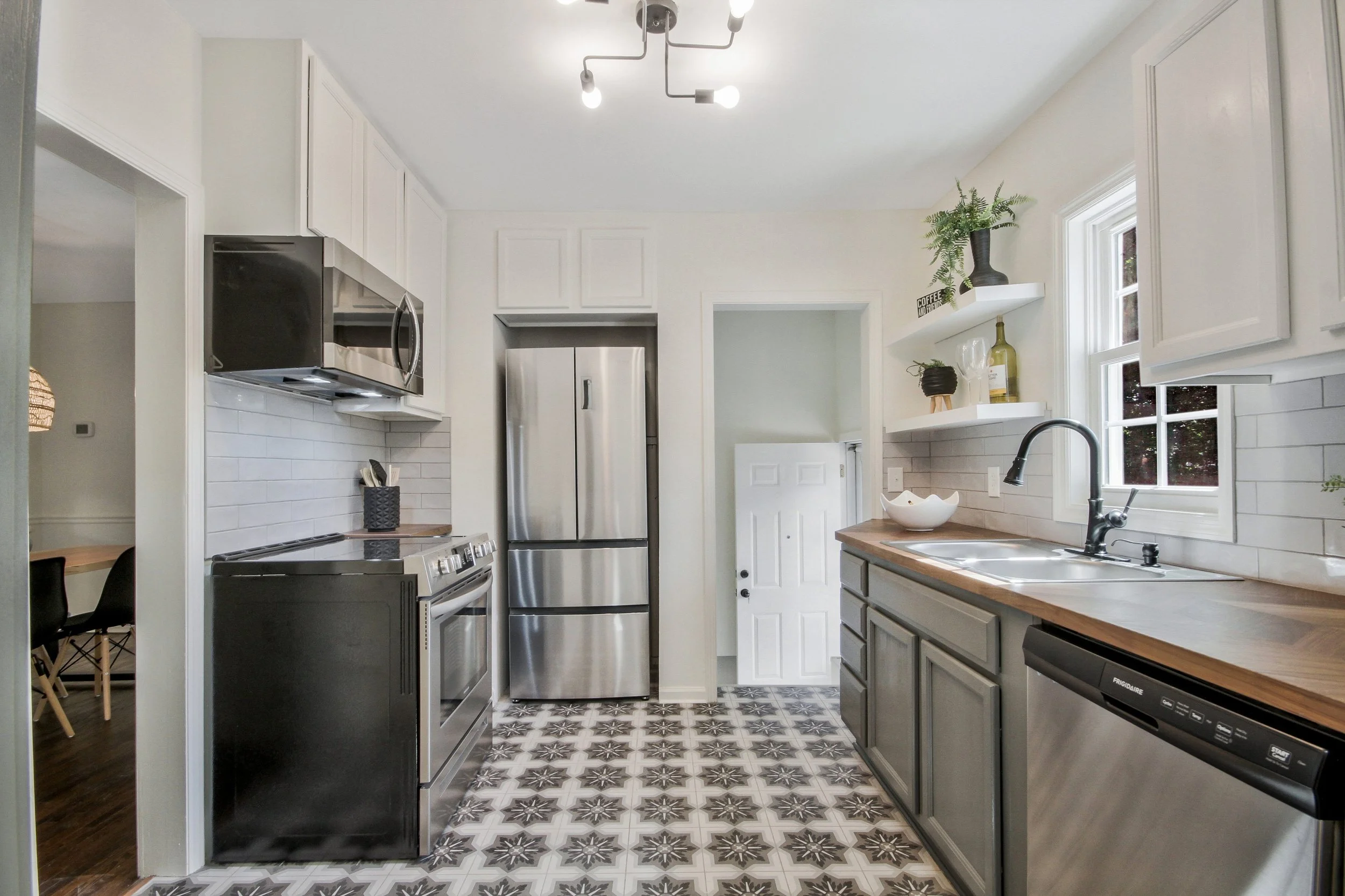 A modern kitchen with white upper cabinets, gray lower cabinets, stainless steel refrigerator, microwave, and oven. The floor has geometric black and white tile. Open shelving with potted plants and wine bottles are above the sink. A window provides natural light.