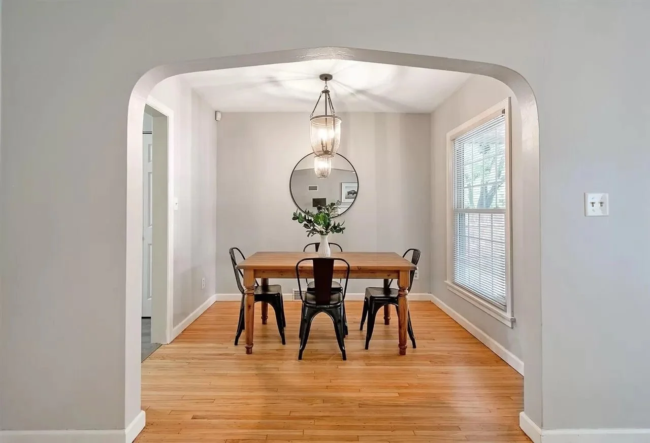 Dining room with a wooden table, four black chairs, a hanging light fixture, a round mirror, a window with blinds, and a vase with greenery.
