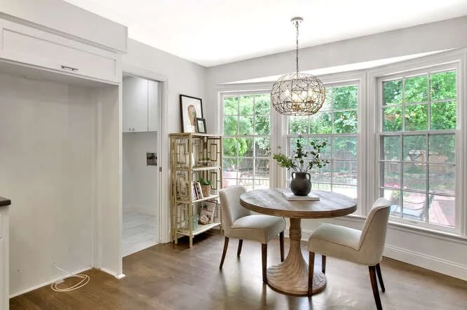 A bright dining area with a round wooden table, four white chairs, a large window with green trees outside, and a modern chandelier hanging from the ceiling.