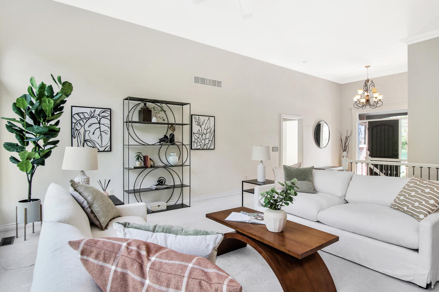 Living room with white sofas, a wooden coffee table, green plants, black metal shelving, framed wall art, lamps, and a chandelier.