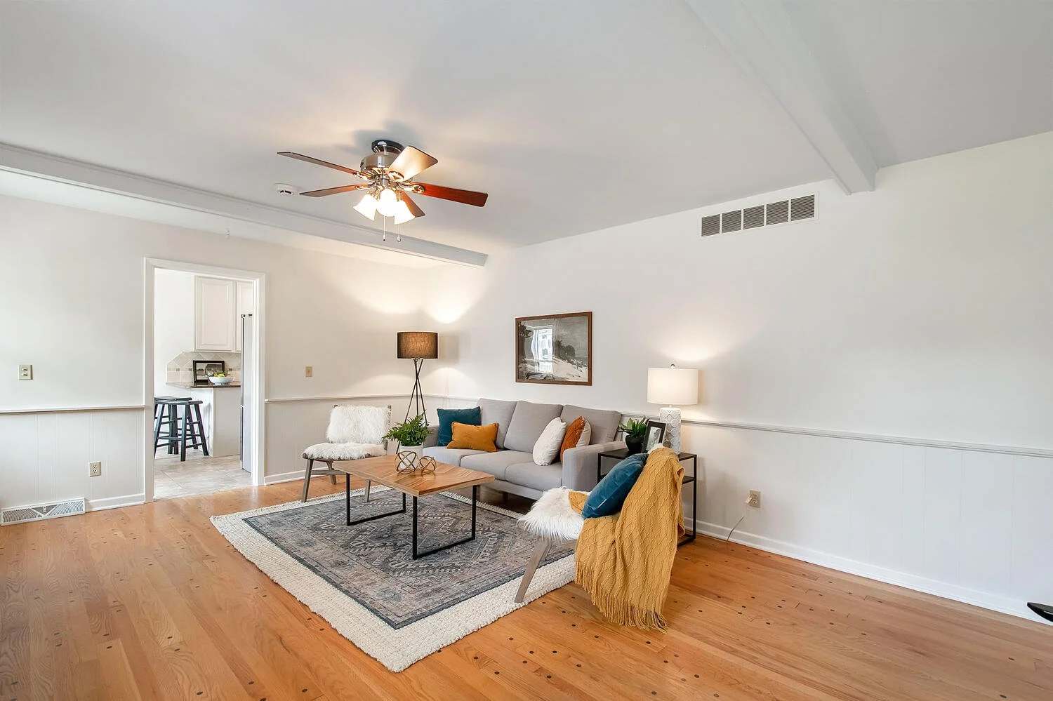 Living room with white walls, hardwood floors, a ceiling fan, a grey sofa with throw pillows, a black side table with a lamp, a yellow chair with a blanket, a small wooden coffee table, a area rug, and a doorway leading to the kitchen.