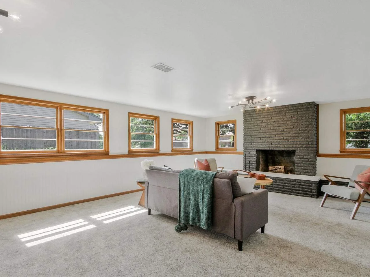 Living room with white walls, four windows with wooden frames, a brick fireplace, a grey sofa with a green throw, armchairs, a round coffee table, and a ceiling light fixture.